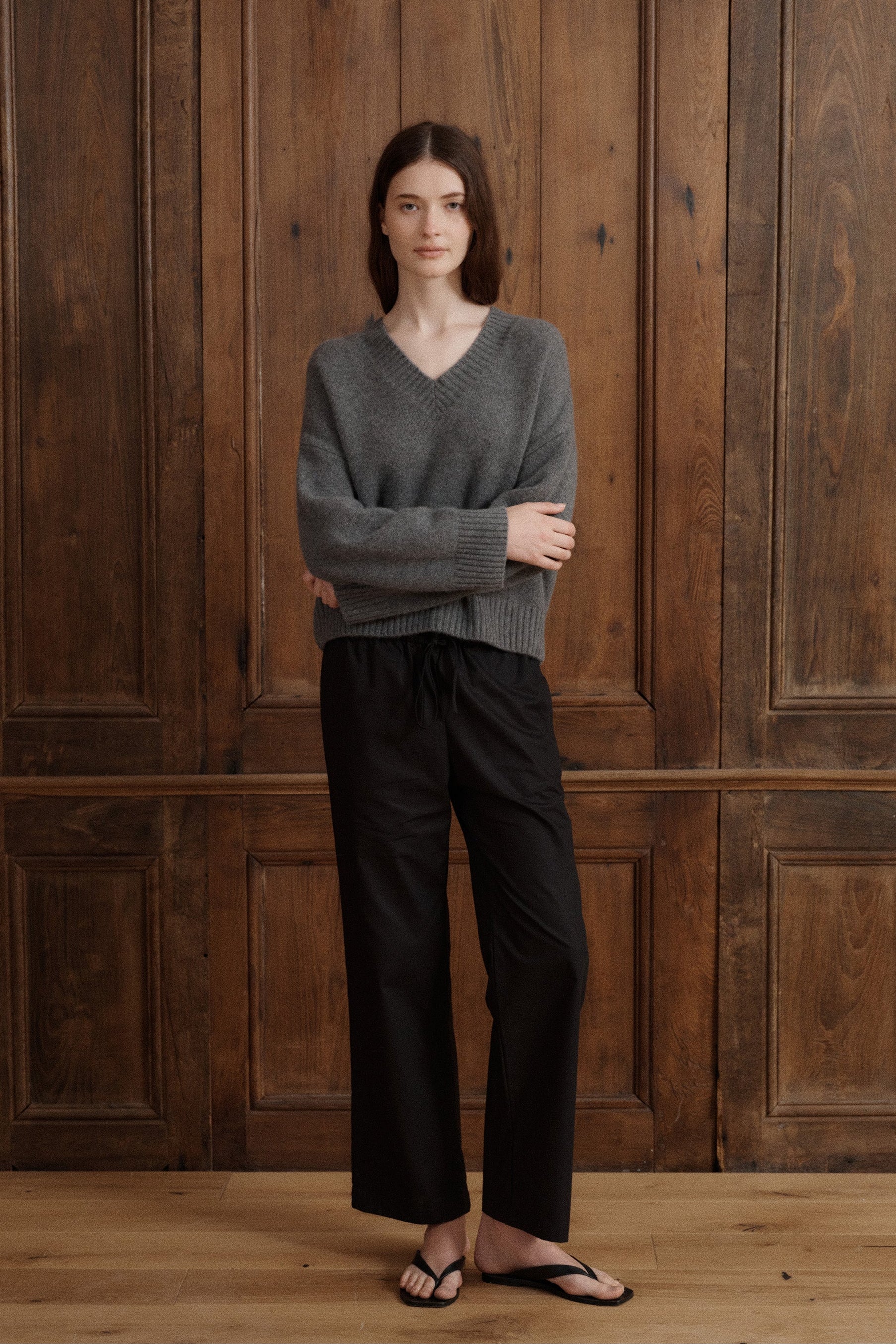 Female model posing wearing a classic V-neck knit sweater in dark grey. Knitted from grade A cashmere with a relaxed fit and dropped shoulders. Featuring wide sleeves and ribbing at the neck, hem, and cuffs. It is paired with black cotton poplin pants and black sandals. - VIR VITA
#color_stonegrey