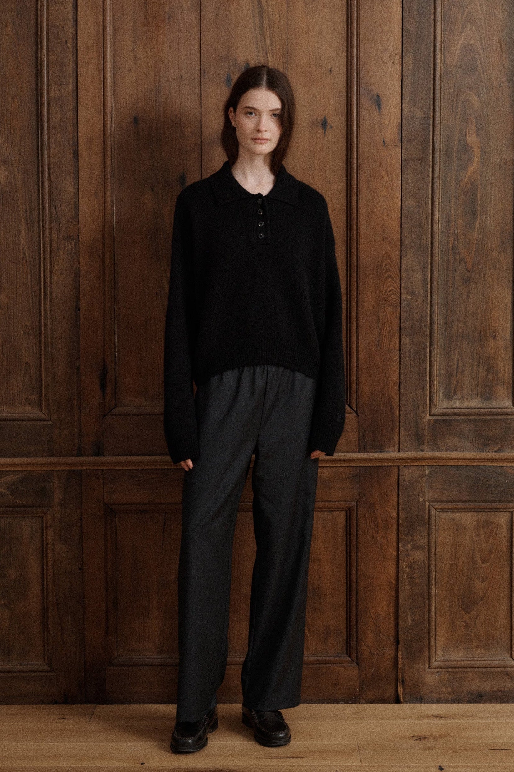 Female model posing wearing a black polo sweater crafted from soft cashmere. Designed with a structured collar, shoulder, and four-button front closure for a timeless look. Paired with dark grey wool elastic waist trousers and black loafers - VIR VITA
#color_black