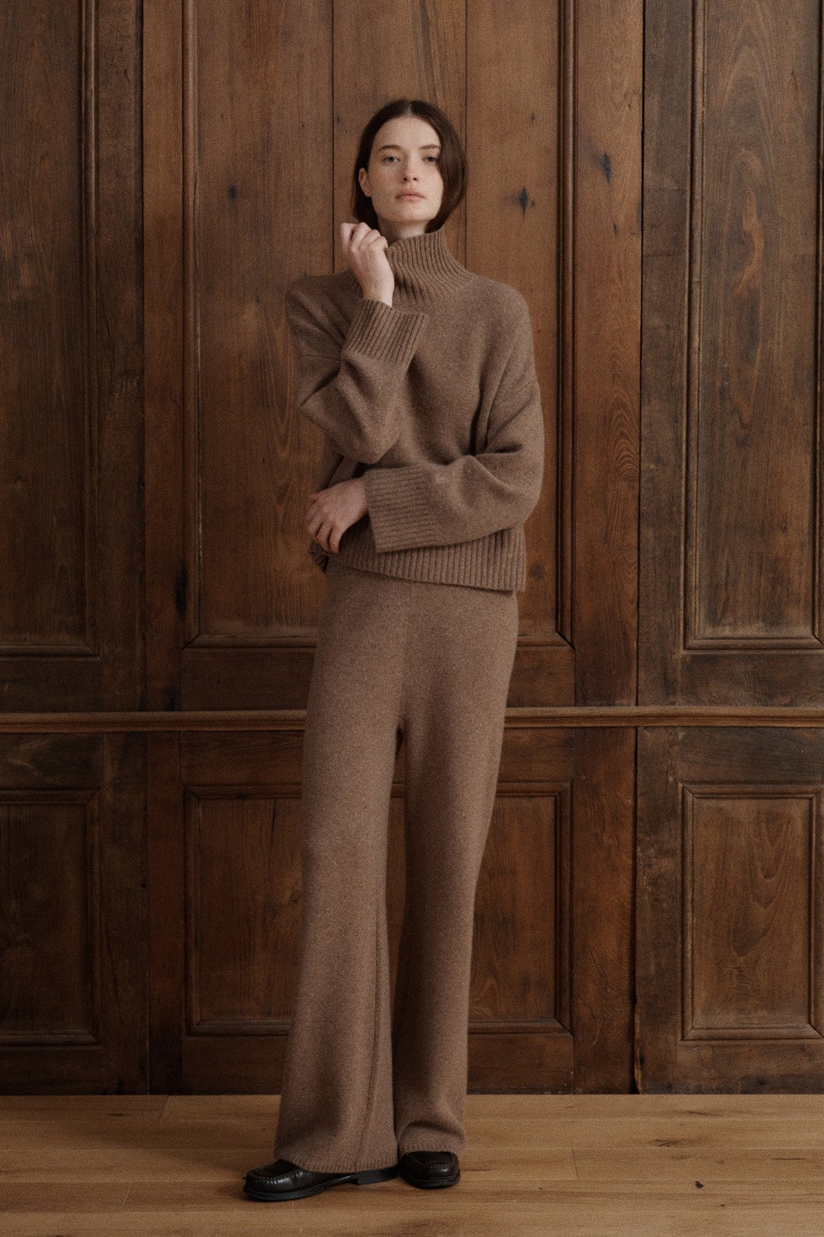 Female model wearing a turtleneck jumper knit in Grade A cashmere in a soft speckled brown color. It features wide balloon sleeves and dropped shoulders for an effortless silhouette. It is paired with matching lounge cashmere pants and black loafers. - VIR VITA
#color_toastedwalnut