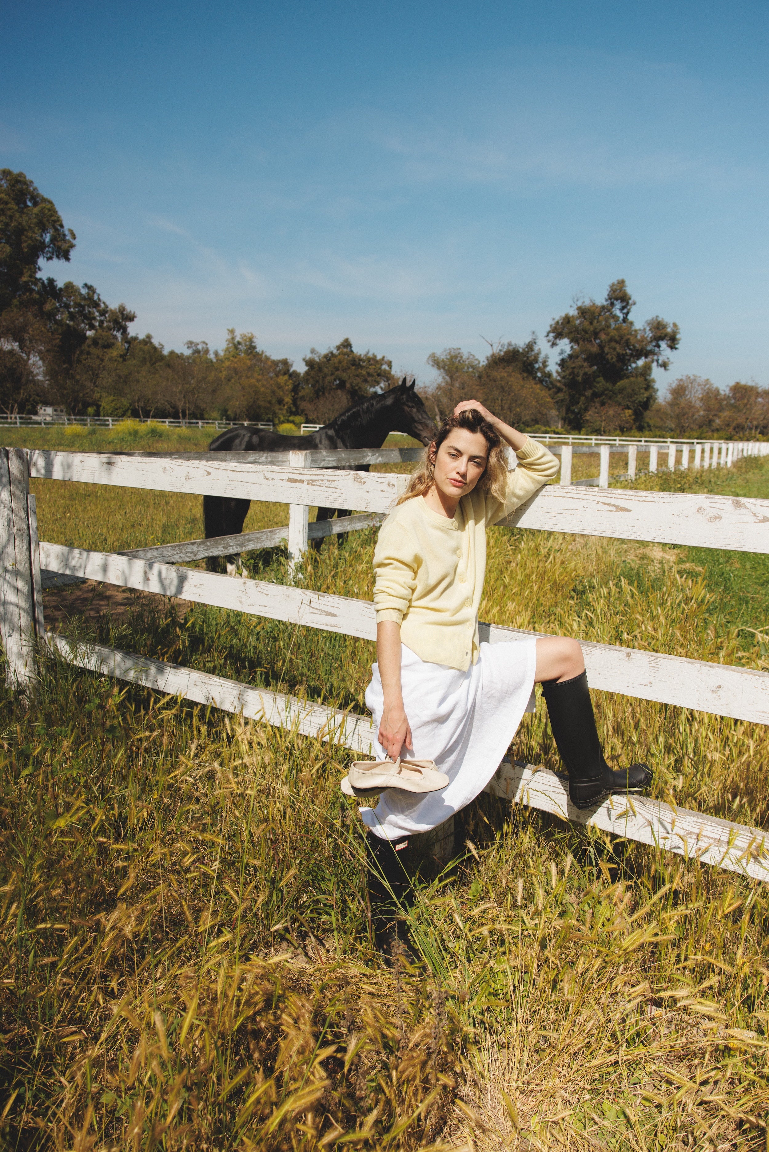 Female model leaning against a fence wearing a pale yellow ultra-soft luxurious cashmere button up cardigan paired with a white linen maxi skirt, and white flats - VIR VITA
#color_butter