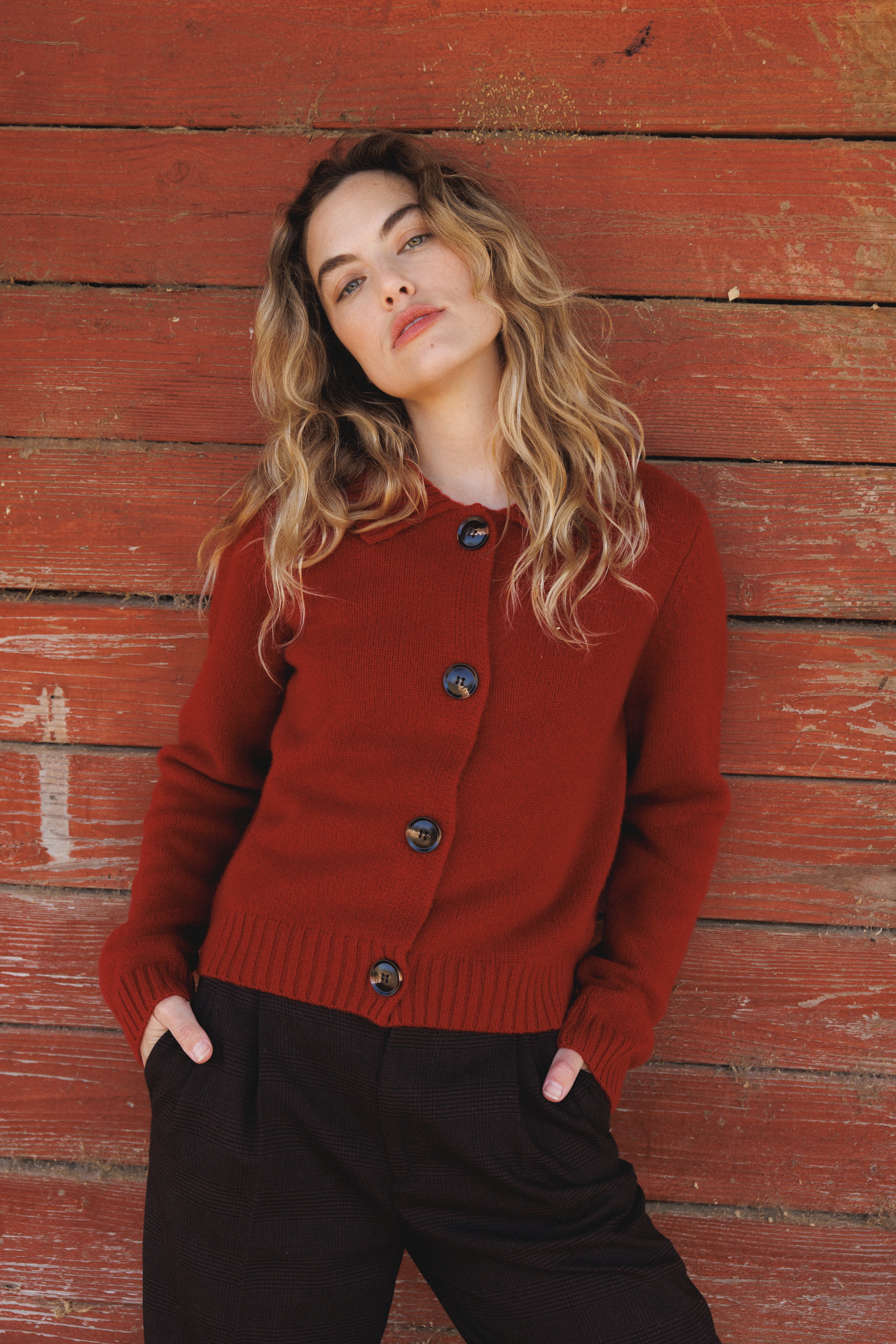 Female model leaning against wall wearing a red ultra-soft luxurious cashmere cardigan featuring a classic silhouette with a collar and large buttons paired with dark brown trousers- VIR VITA
#color_cherry