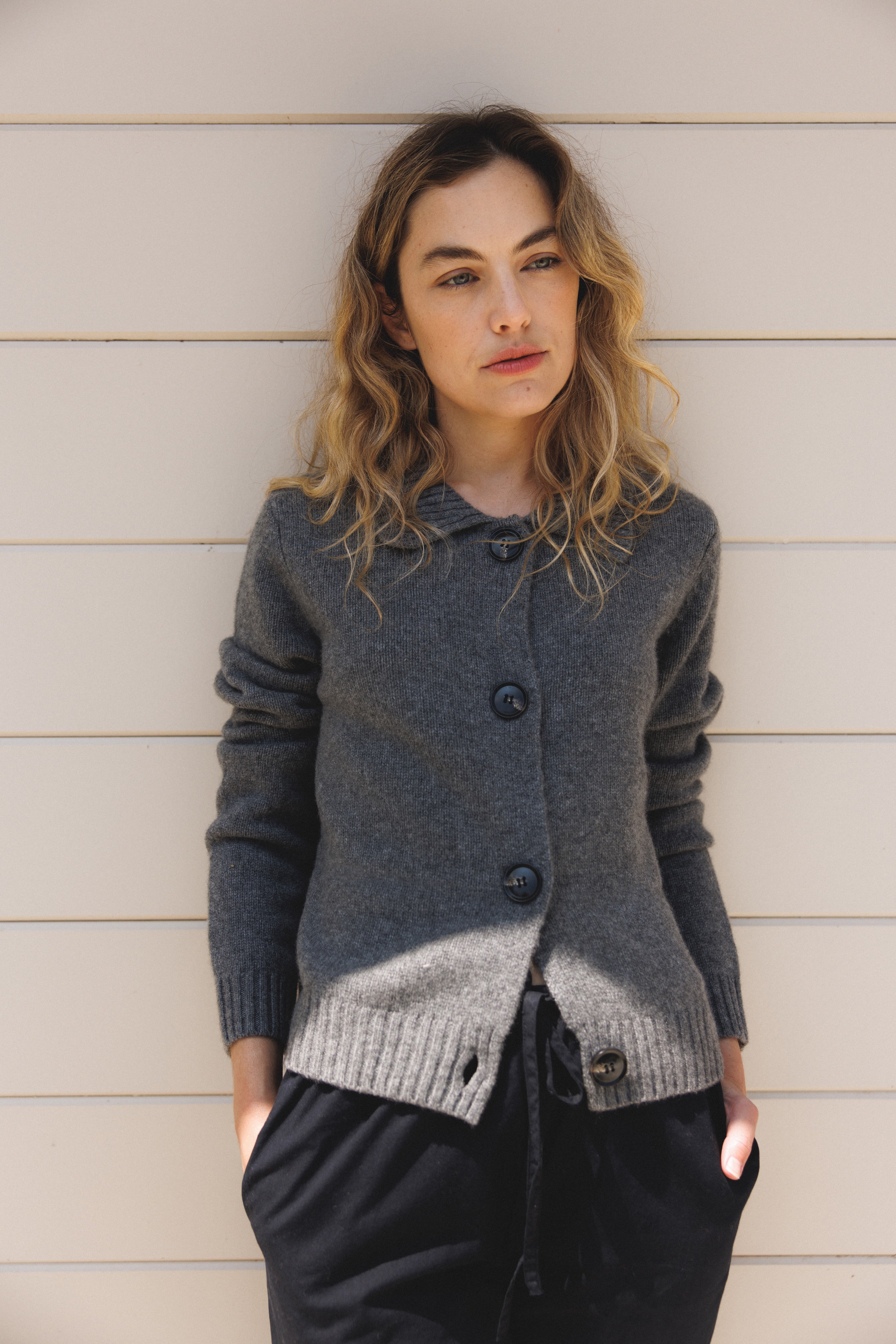 Female model wearing a grey ultra-soft luxurious cashmere cardigan featuring a classic silhouette with a collar and large buttons paired with black drawstring pants- VIR VITA
#color_stone grey