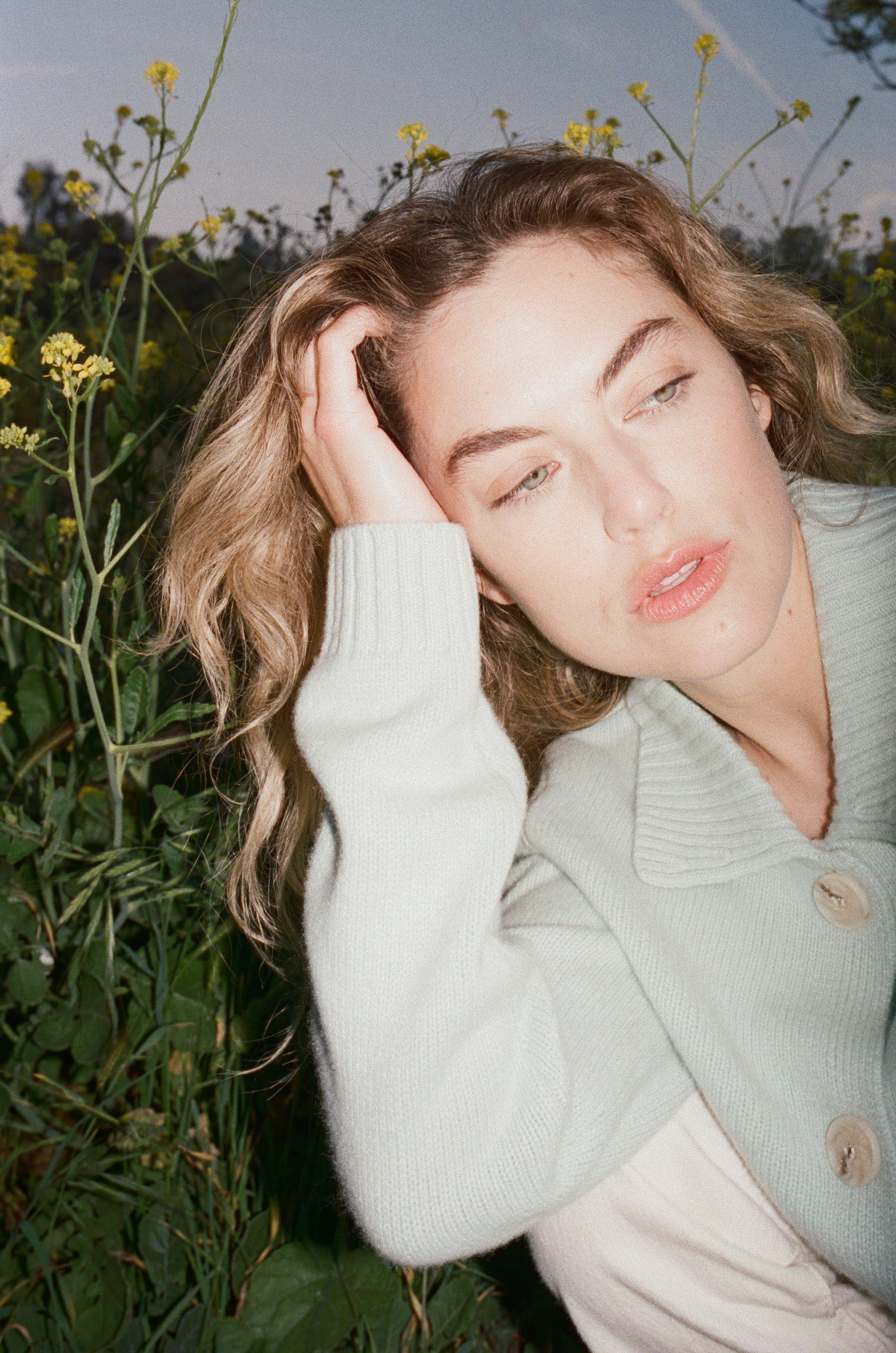 Female model sitting in grass wearing a teal blue ultra-soft luxurious cashmere cardigan featuring a classic silhouette with a collar and large buttons paired with white jeans- VIR VITA
#color_sea glass