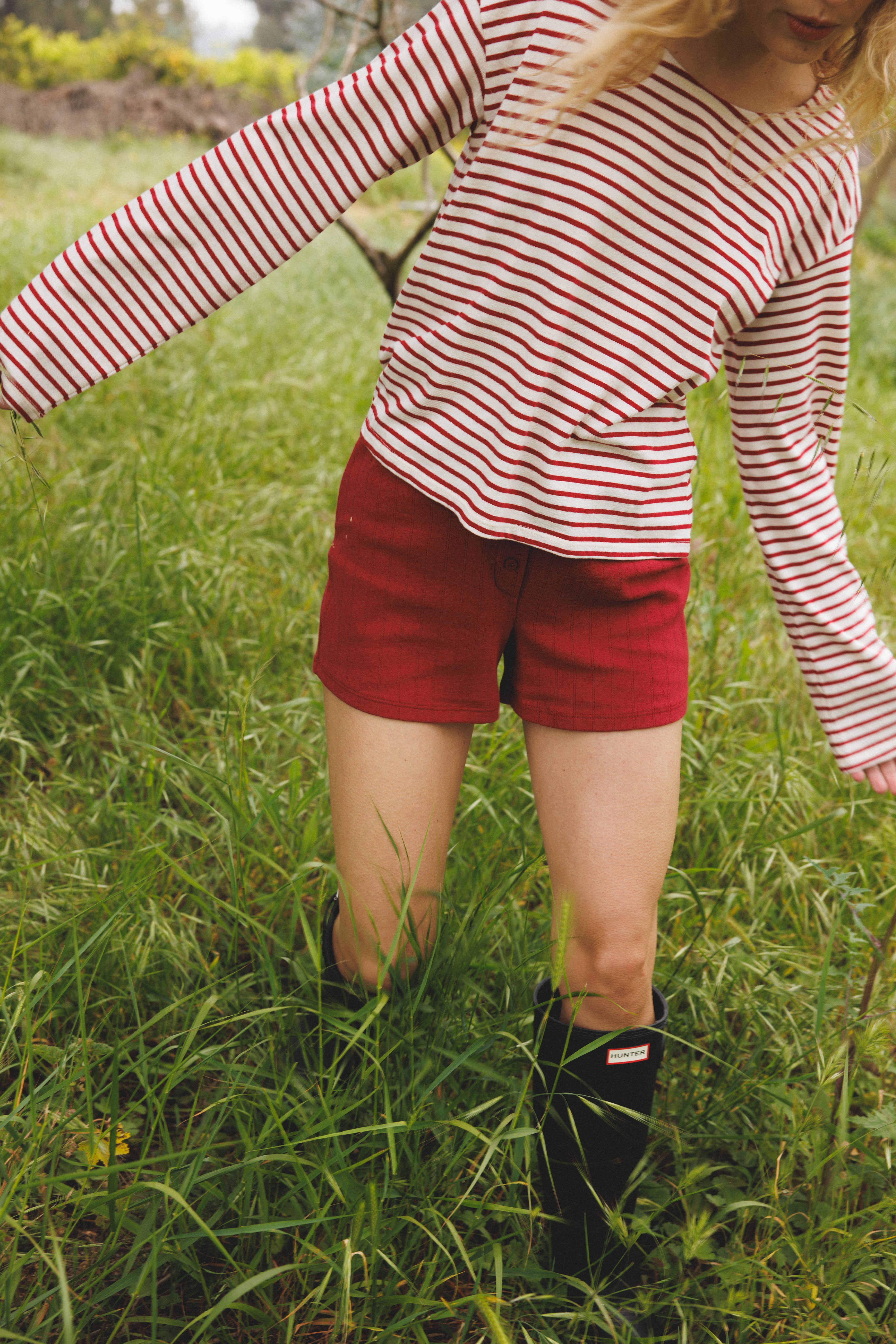 Female model walking through grass wearing a sustainable long sleeve striped red and white top featuring a flattering neckline and oversized fit paired with pointelle shorts in red. - VIR VITA
#color_classic red stripe