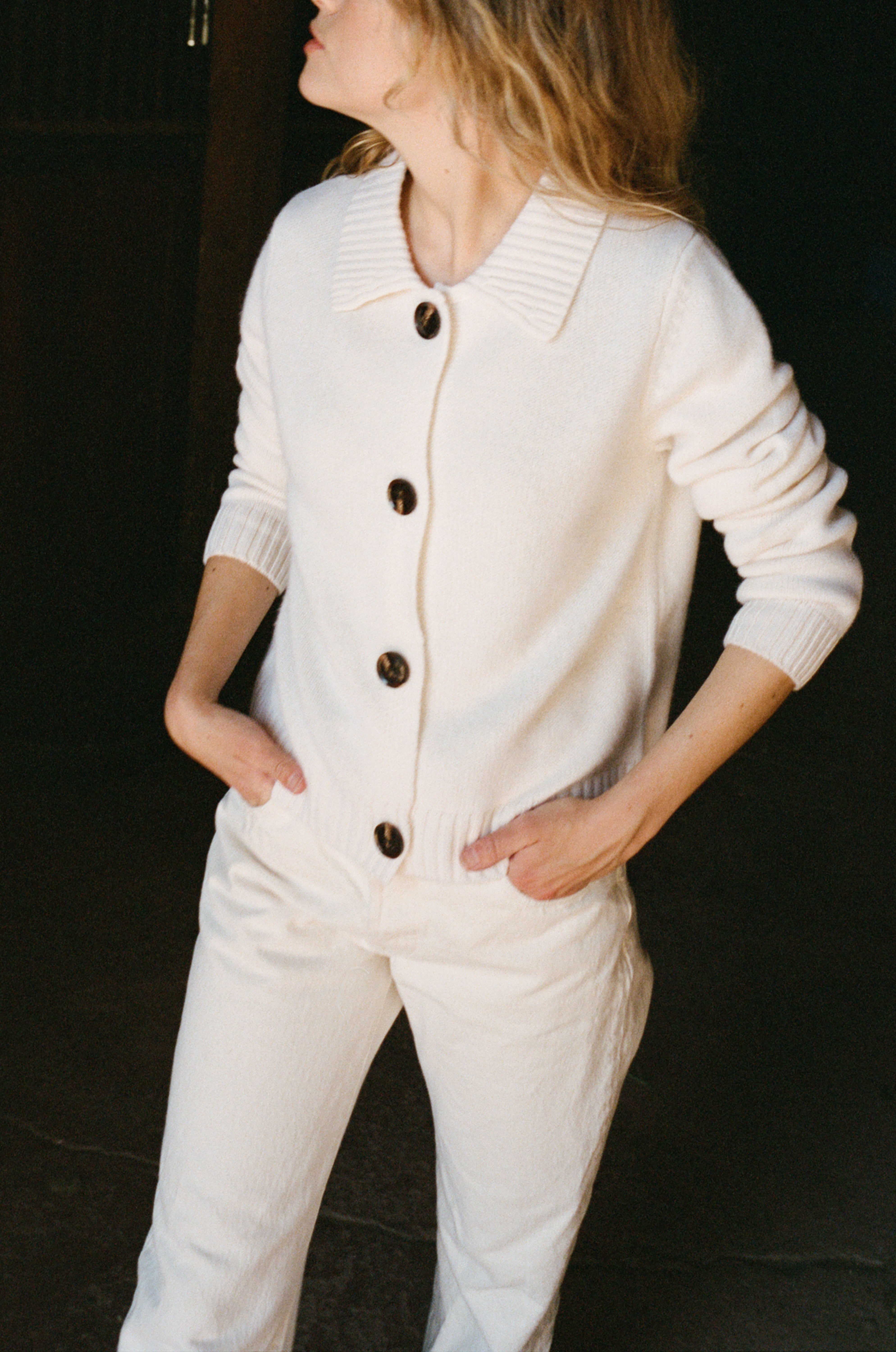 Female model wearing a white ultra-soft luxurious cashmere cardigan featuring a classic silhouette with a collar and large buttons paired with white jeans and brown boots - VIR VITA
#color_bone