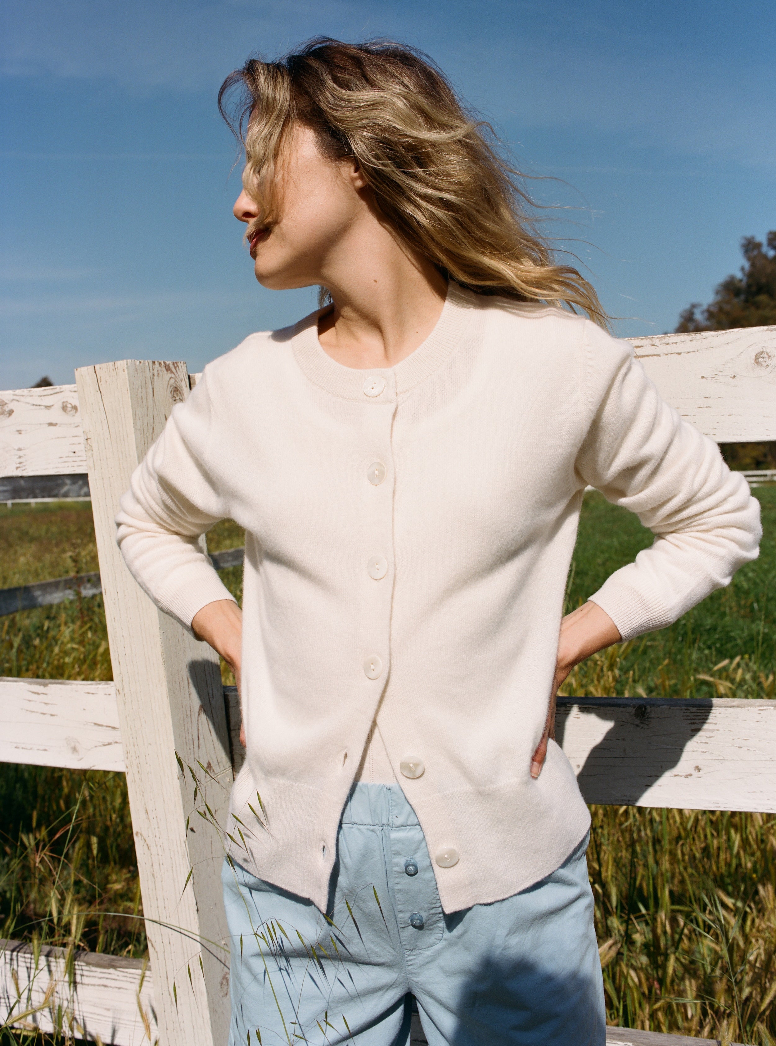 Female model standing next to a fence wearing a white ultra-soft luxurious cashmere button up cardigan paired with light blue cotton poplin pants - VIR VITA
#color_bone