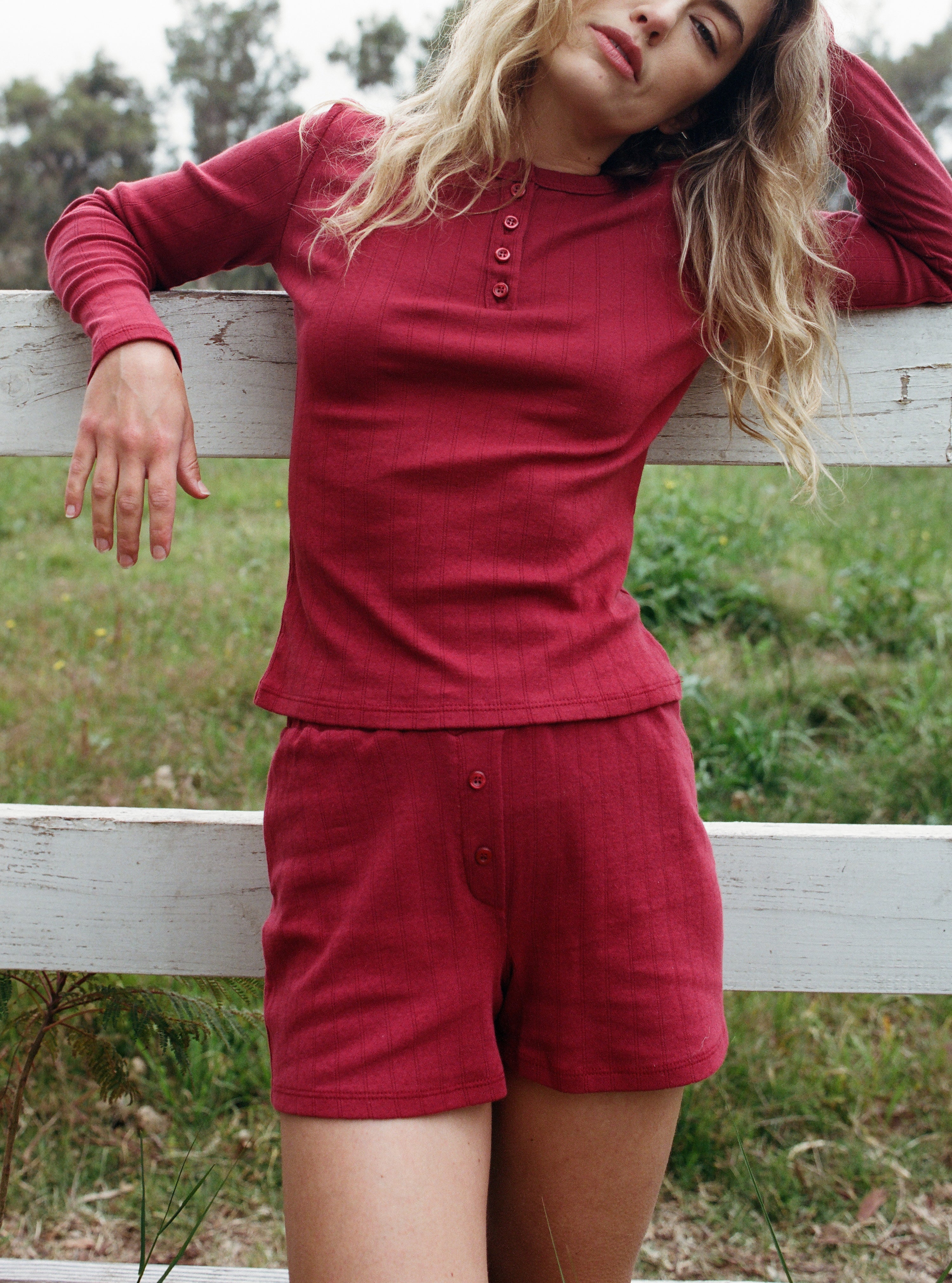 Female model leaning against a fence wearing a dark red cotton pointelle henley long sleeve top with matching buttons paired with cotton pointelle lounge shorts in dark red - VIR VITA
#color_sun dried tomato