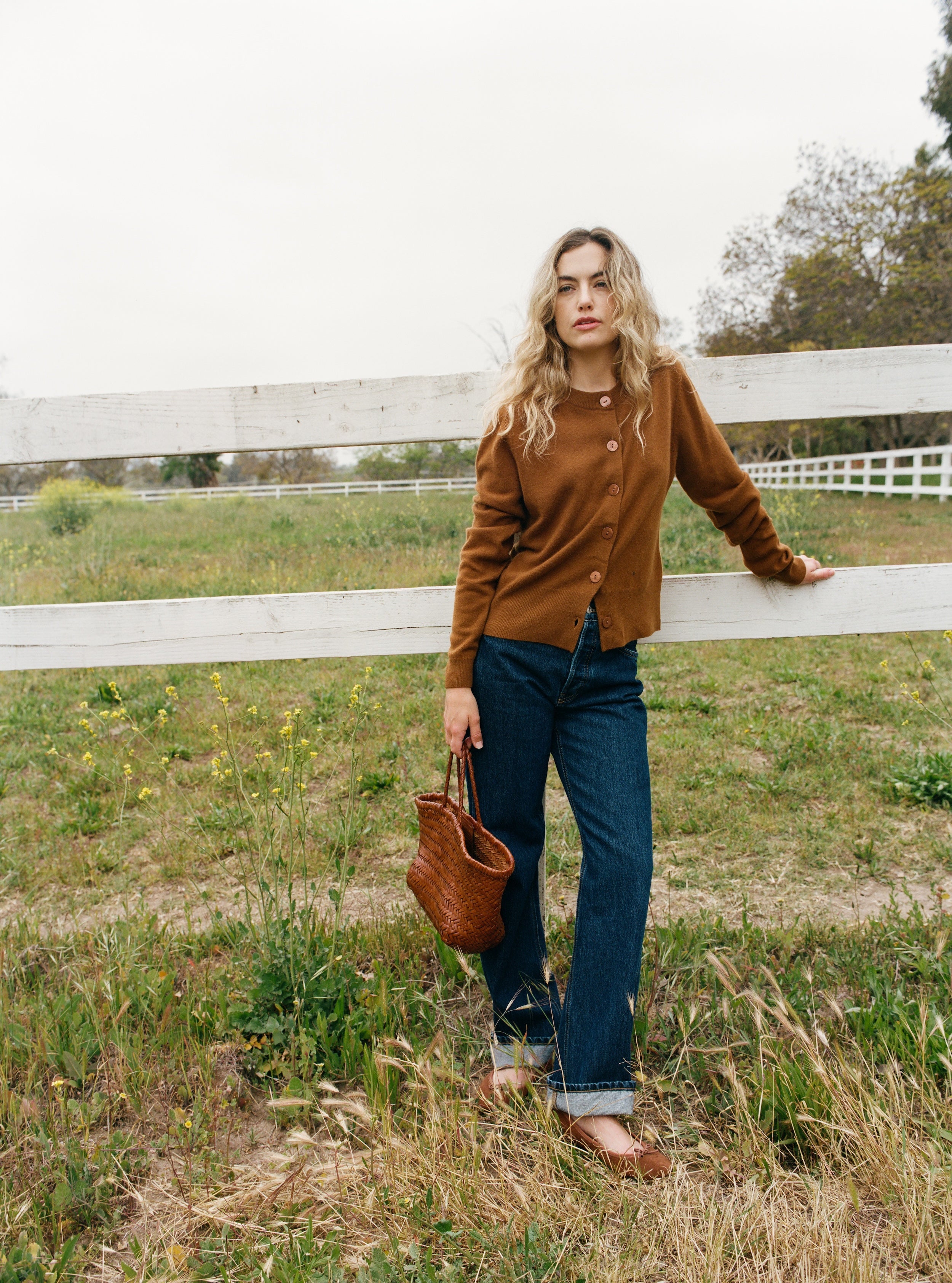 Female model standing next to a fence wearing a brown ultra-soft luxurious cashmere button up cardigan paired with blue jeans, brown flats, and a brown woven bag - VIR VITA
#color_fawn