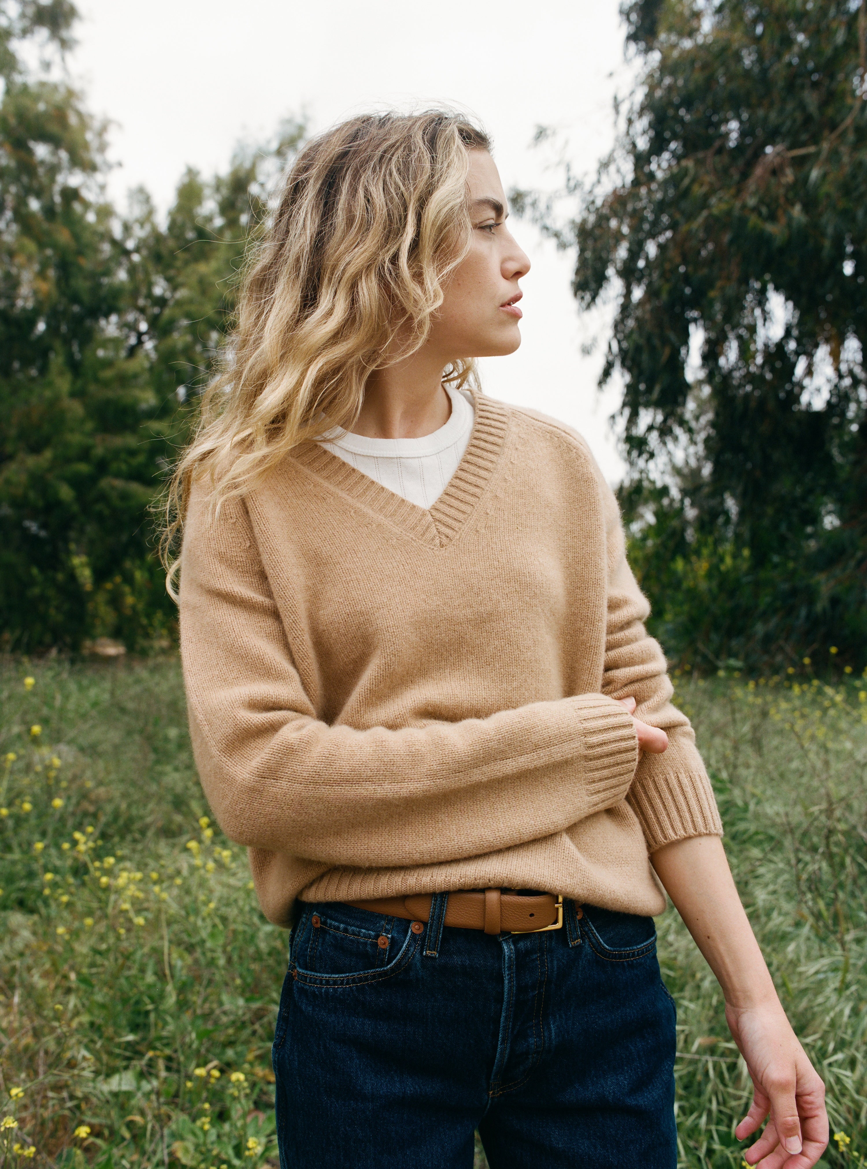 Female model standing in a field wearing an ultra-soft luxurious cashmere pullover sweater featuring a v-neck design in a light brown color paired with dark blue jeans, a brown belt, and a white pointelle tee - VIR VITA
#color_desert