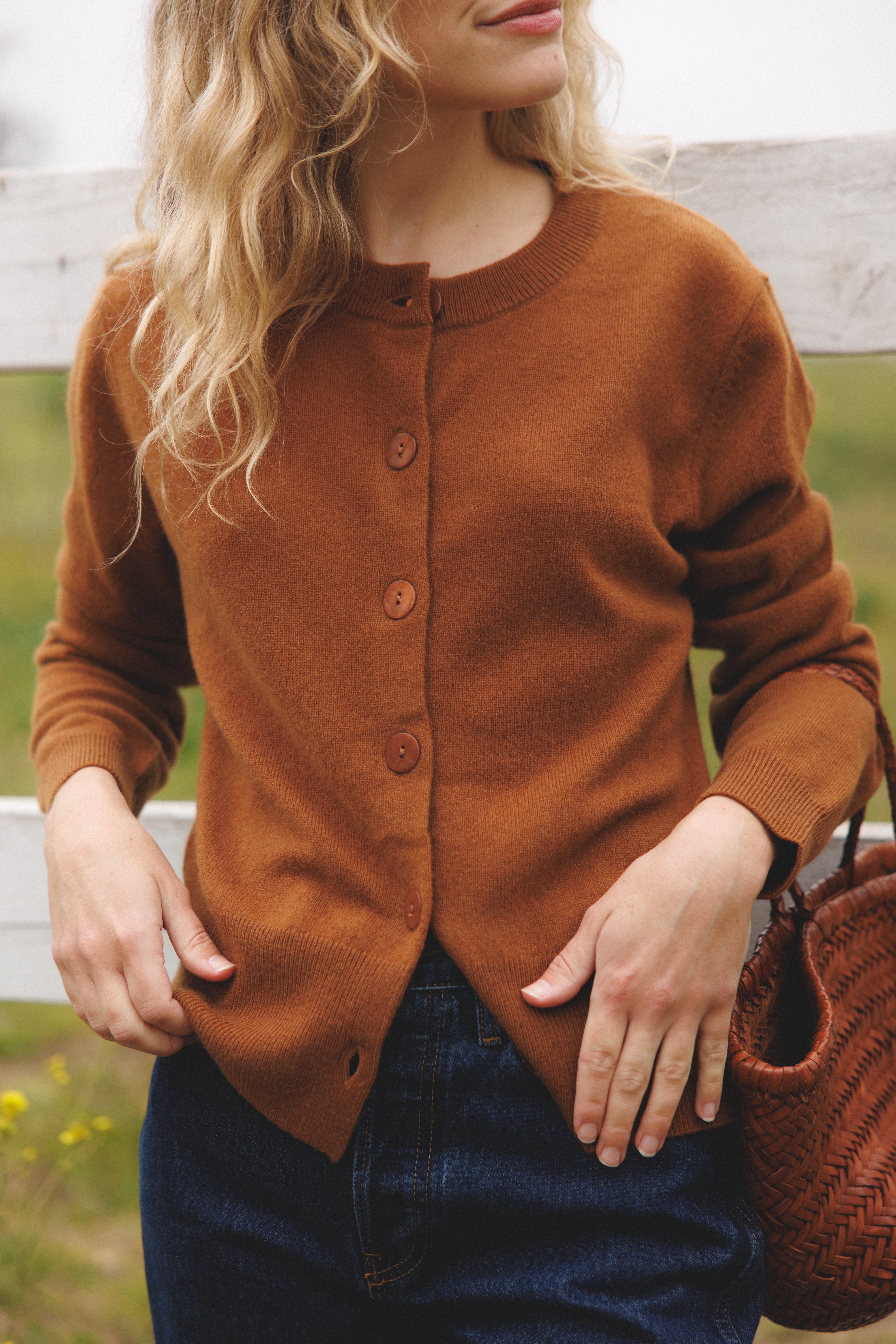Female model leaning against a fence wearing a brown ultra-soft luxurious cashmere button up cardigan paired with blue jeans and a brown woven bag - VIR VITA
#color_fawn