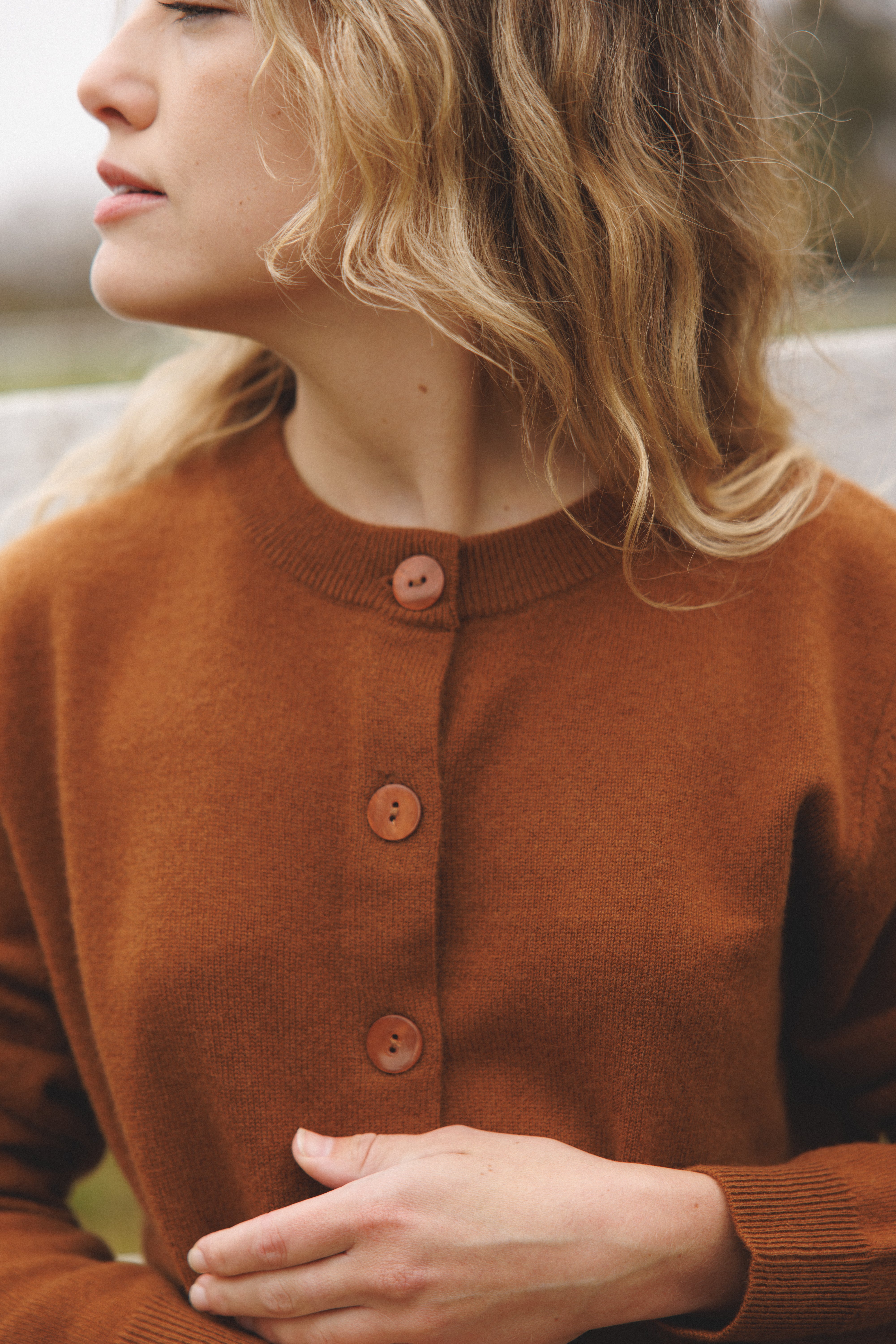 Female model leaning against a fence wearing a brown ultra-soft luxurious cashmere button up cardigan paired with blue jeans and a brown woven bag - VIR VITA
#color_fawn