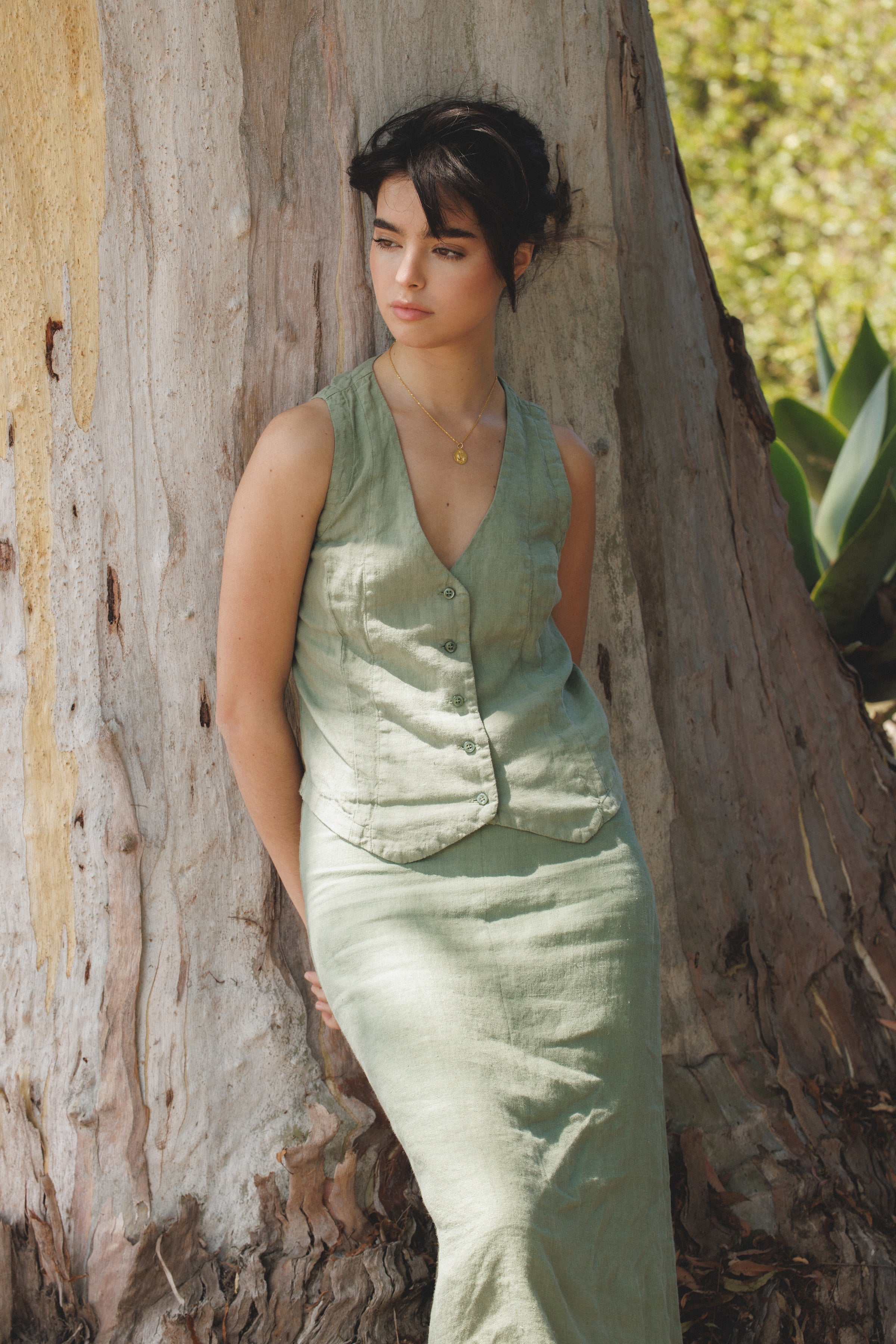 Female model wearing the premium 100% linen maxi skirt featuring a high waisted silhouette in sage green paired with a linen button up vest in sage green next to a tree- VIR VITA
#color_sage