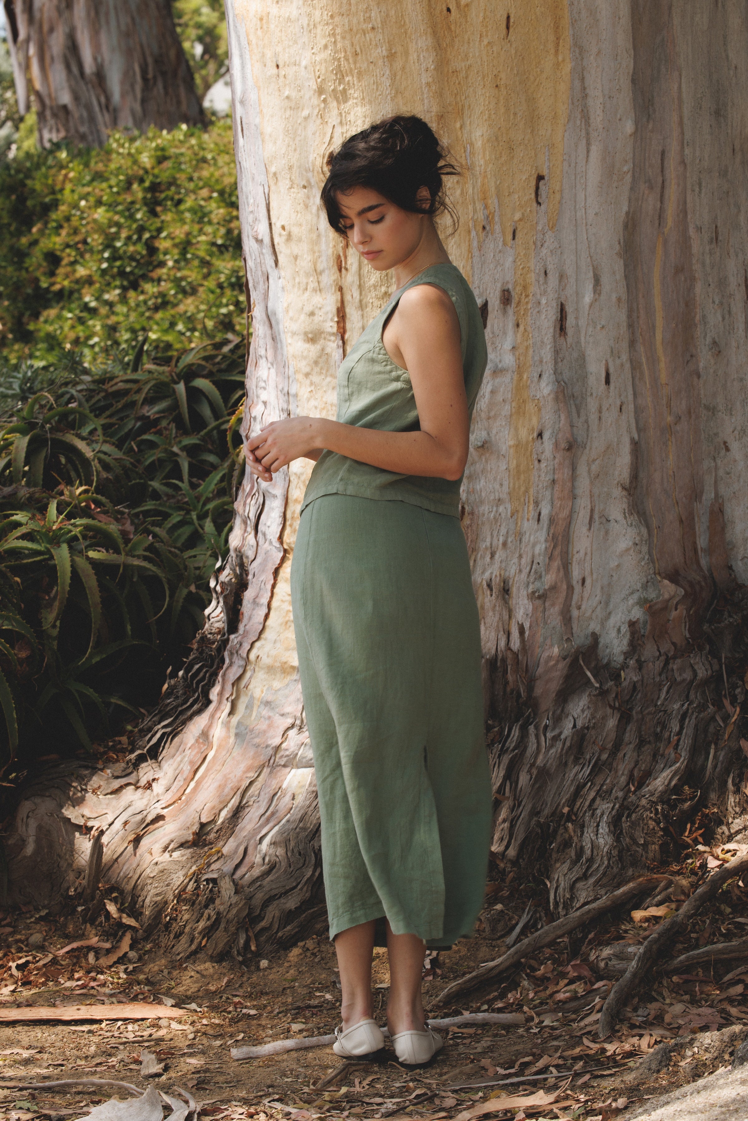 Female model wearing the premium 100% linen maxi skirt featuring a high waisted silhouette in sage green paired with a linen button up vest in sage green with white flats next to a tree- VIR VITA
#color_sage