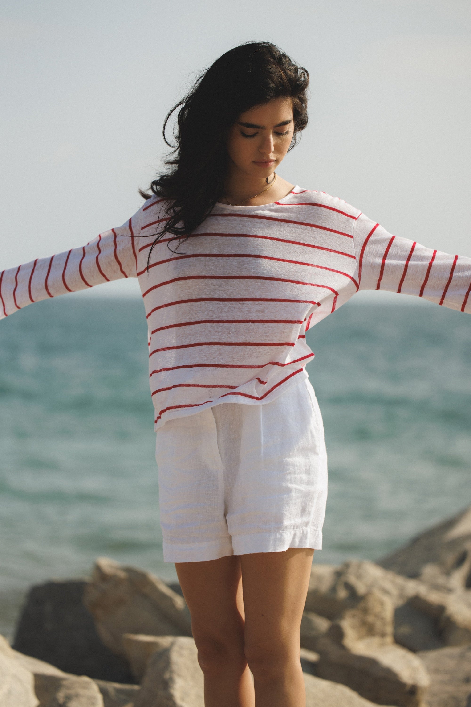 Female model wearing a 100% linen relaxed short featuring double pleats in white paired with a long sleeve linen striped top in white/red on the beach- VIR VITA
#color_white