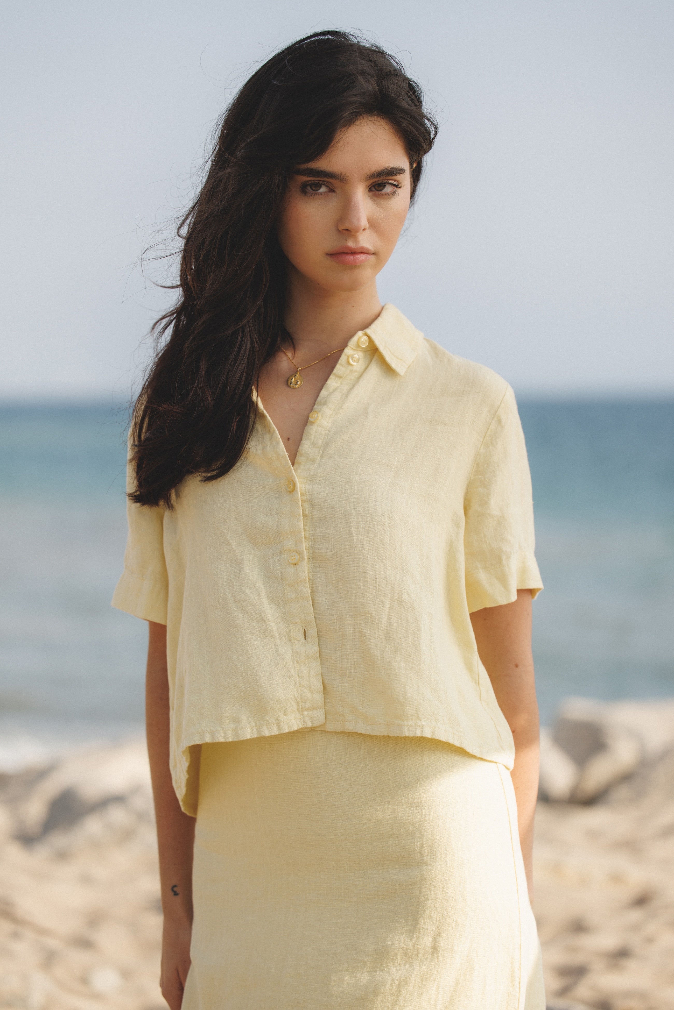Female model wearing a premium 100% linen maxi skirt featuring a high waisted silhouette in pale yellow paired with a linen button up short sleeve shirt in pale yellow on the beach- VIR VITA
#color_butter