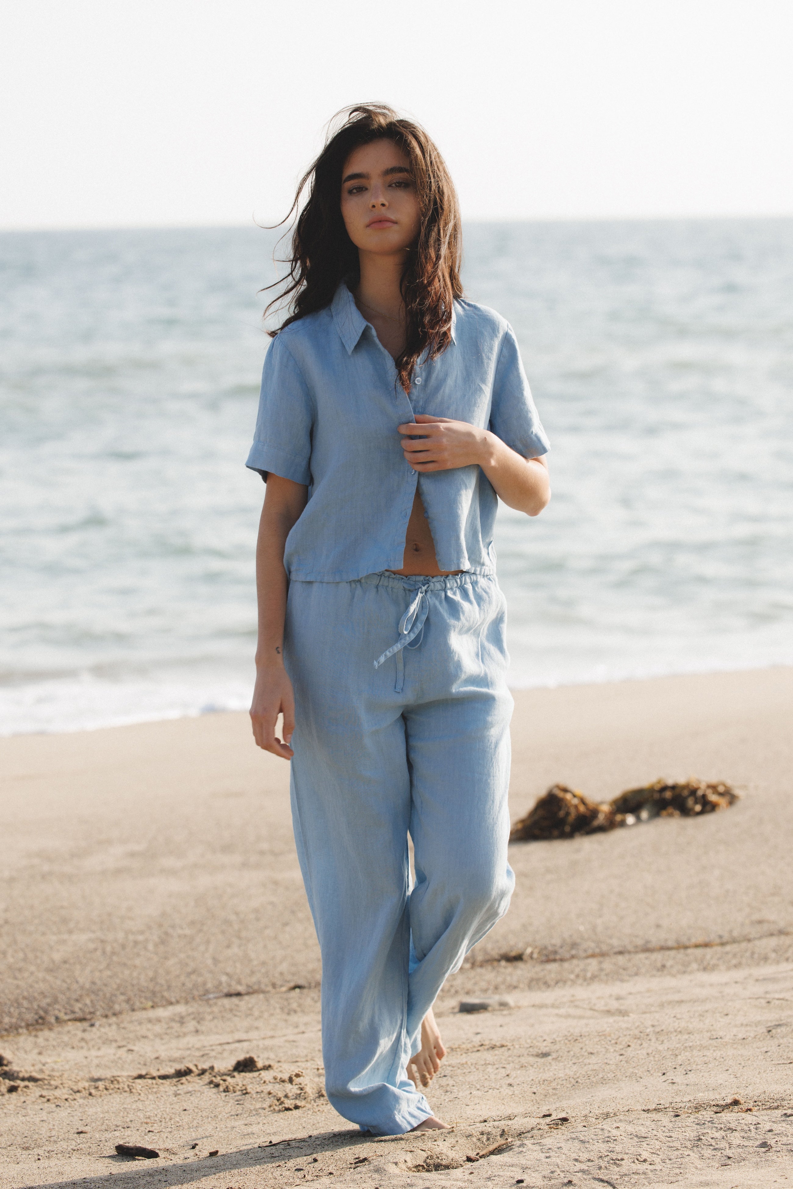 Female model wearing sustainable light blue button up short sleeve 100% linen shirt and matching drawstring linen pant at the beach - VIR VITA
#color_baby blue