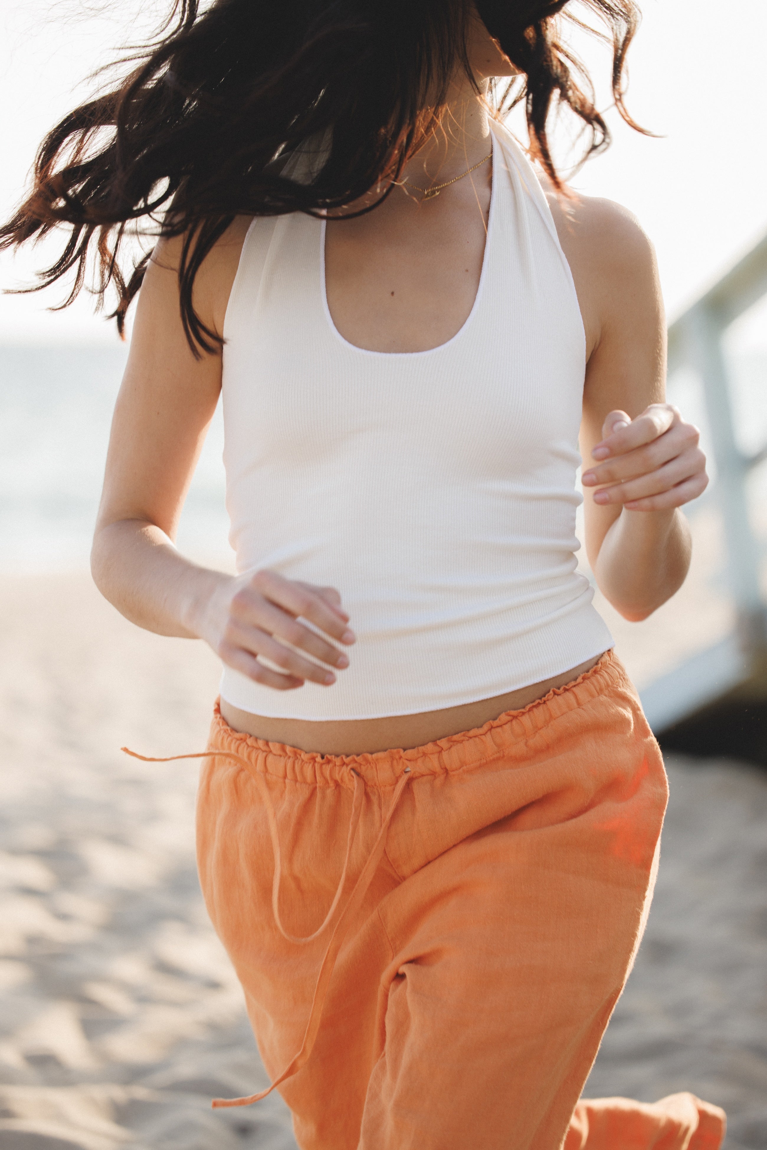 Female model wearing sustainable orange drawstring 100% linen pants featuring a straight leg fit and white modal halter top at the beach - VIR VITA
#color_melon