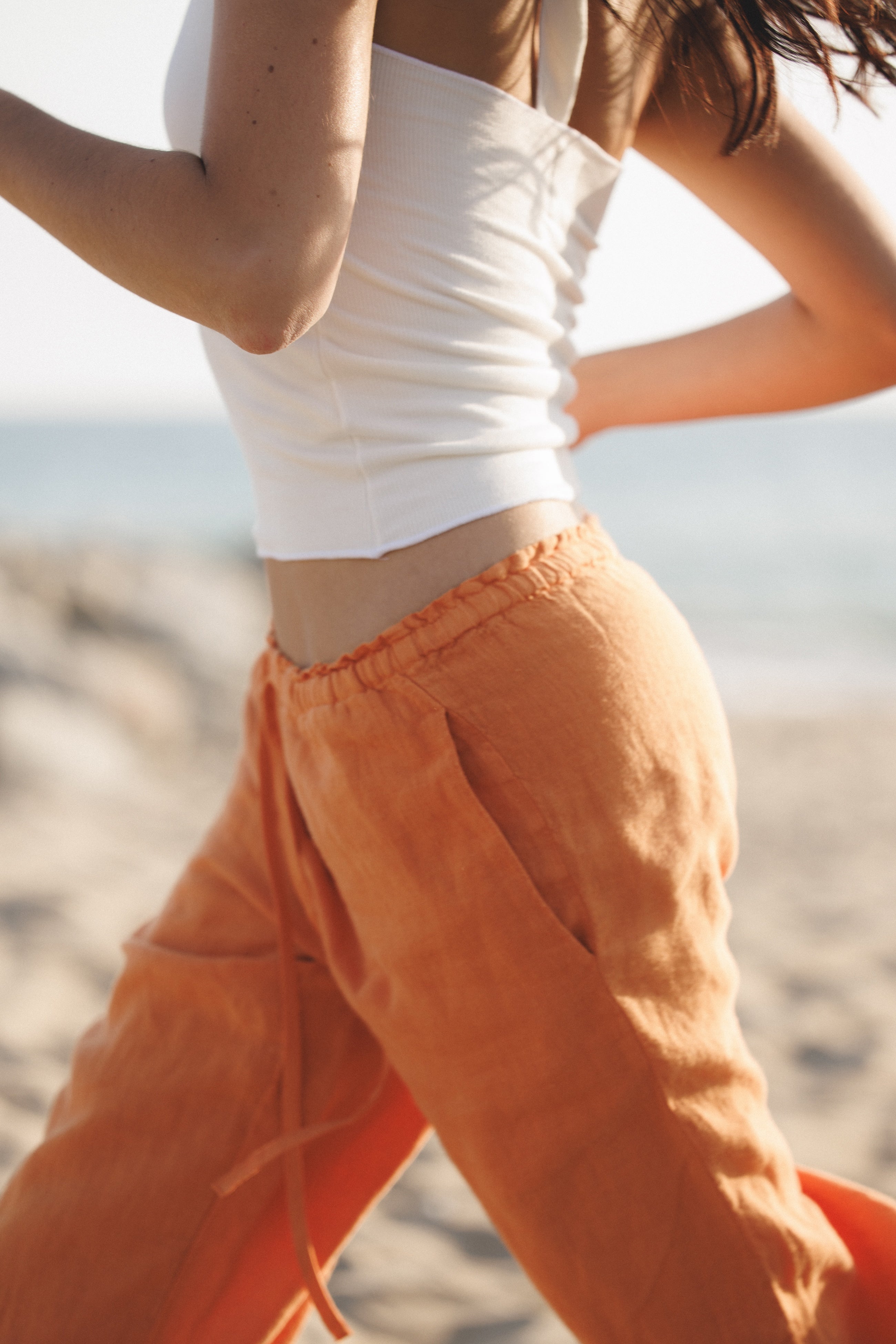 Female model wearing sustainable orange drawstring 100% linen pants featuring a straight leg fit and white modal halter top at the beach - VIR VITA
#color_melon
