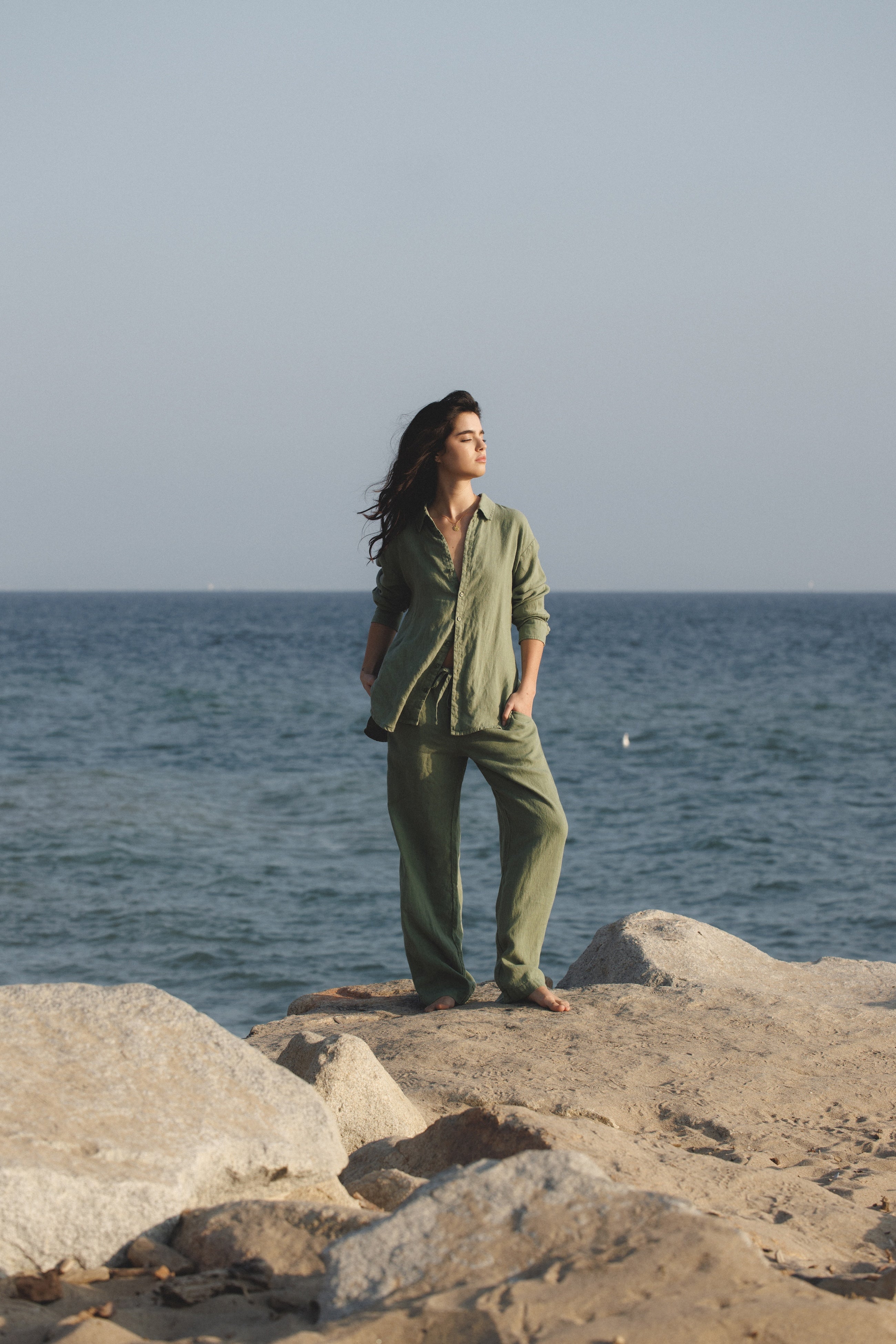 Female model wearing sustainable sage green drawstring 100% linen pants featuring a straight leg fit and sage green long sleeve button up linen shirt at the beach - VIR VITA
#color_sage