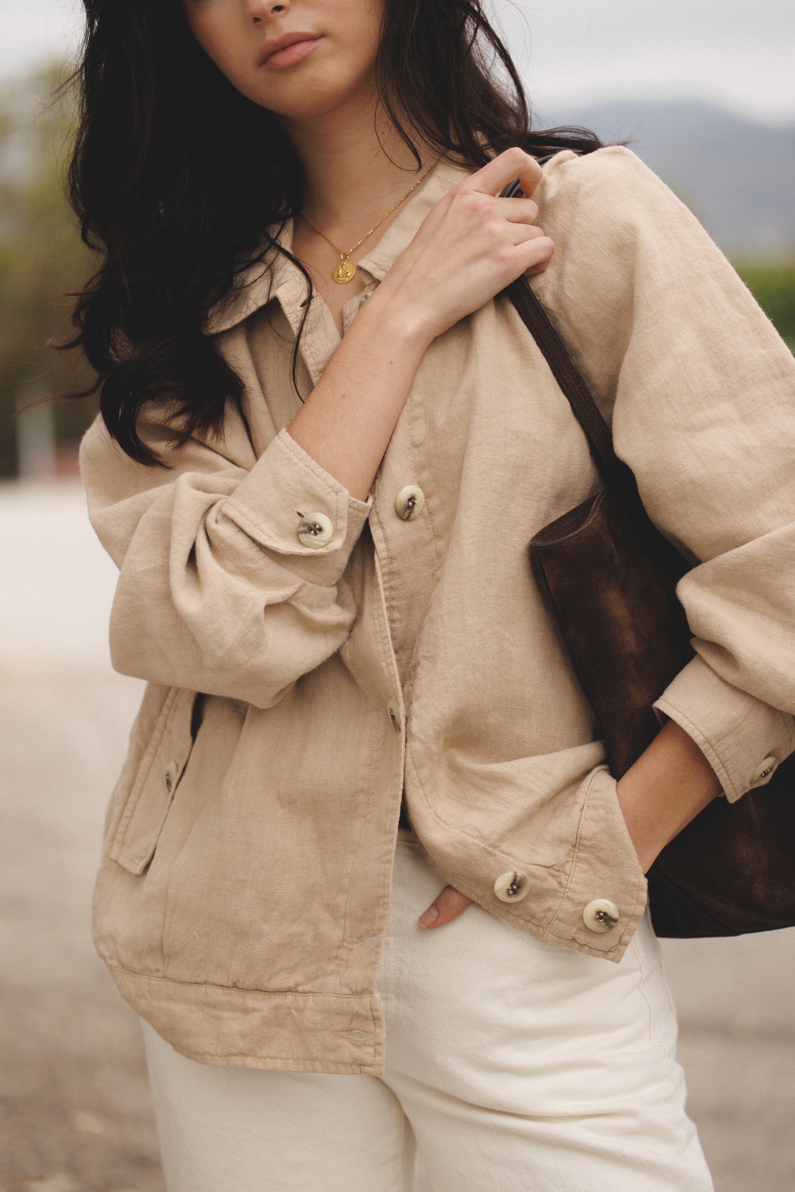 Female model wearing sustainable toffee beige 100% linen barn jacket holding a brown suede bag and wearing white jeans - VIR VITA
#color_toffee