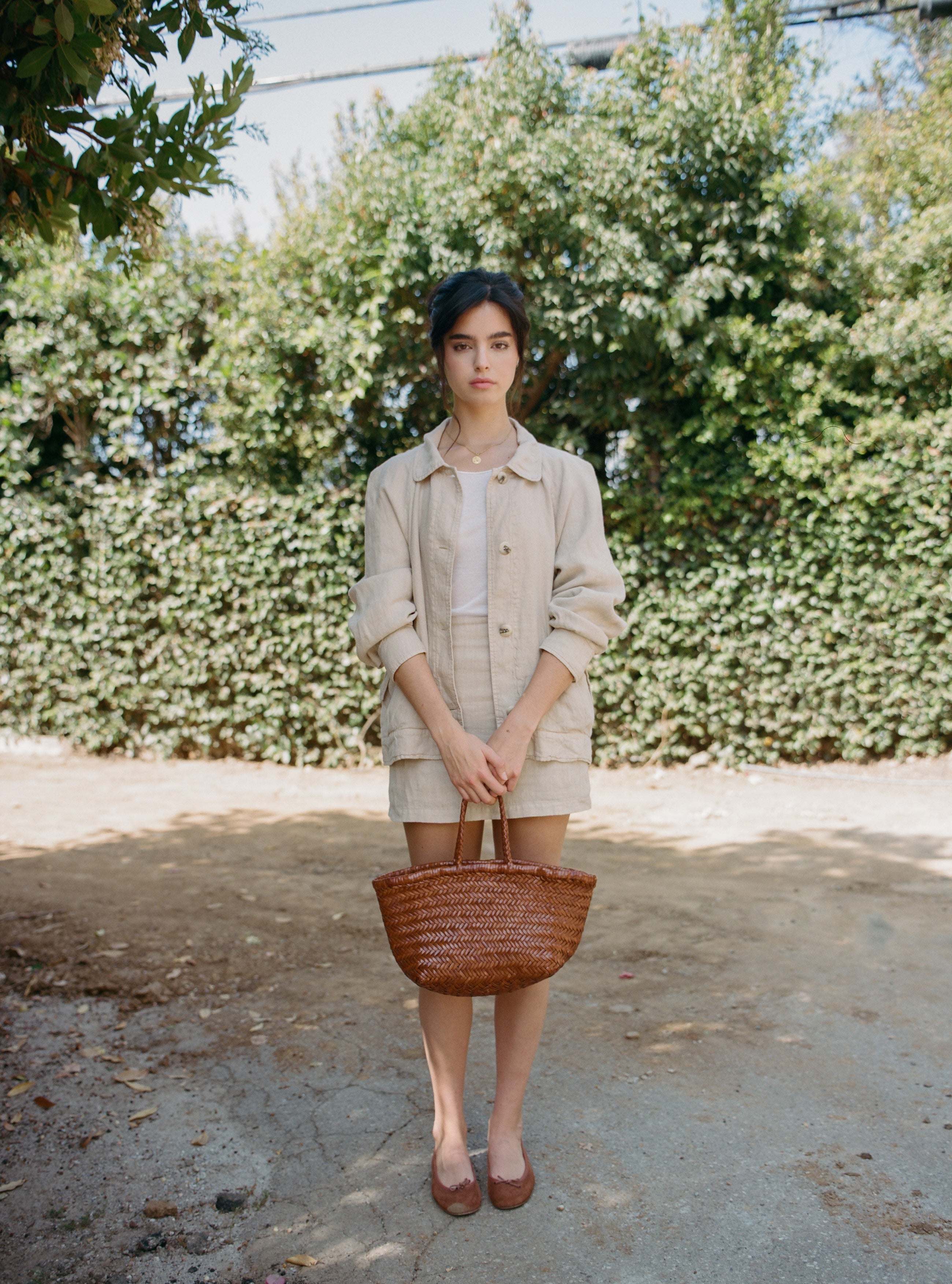 Female model wearing a sustainable beige 100% linen barn jacket and matching beige linen mini skirt holding a leather basket bag- VIR VITA
#color_beige