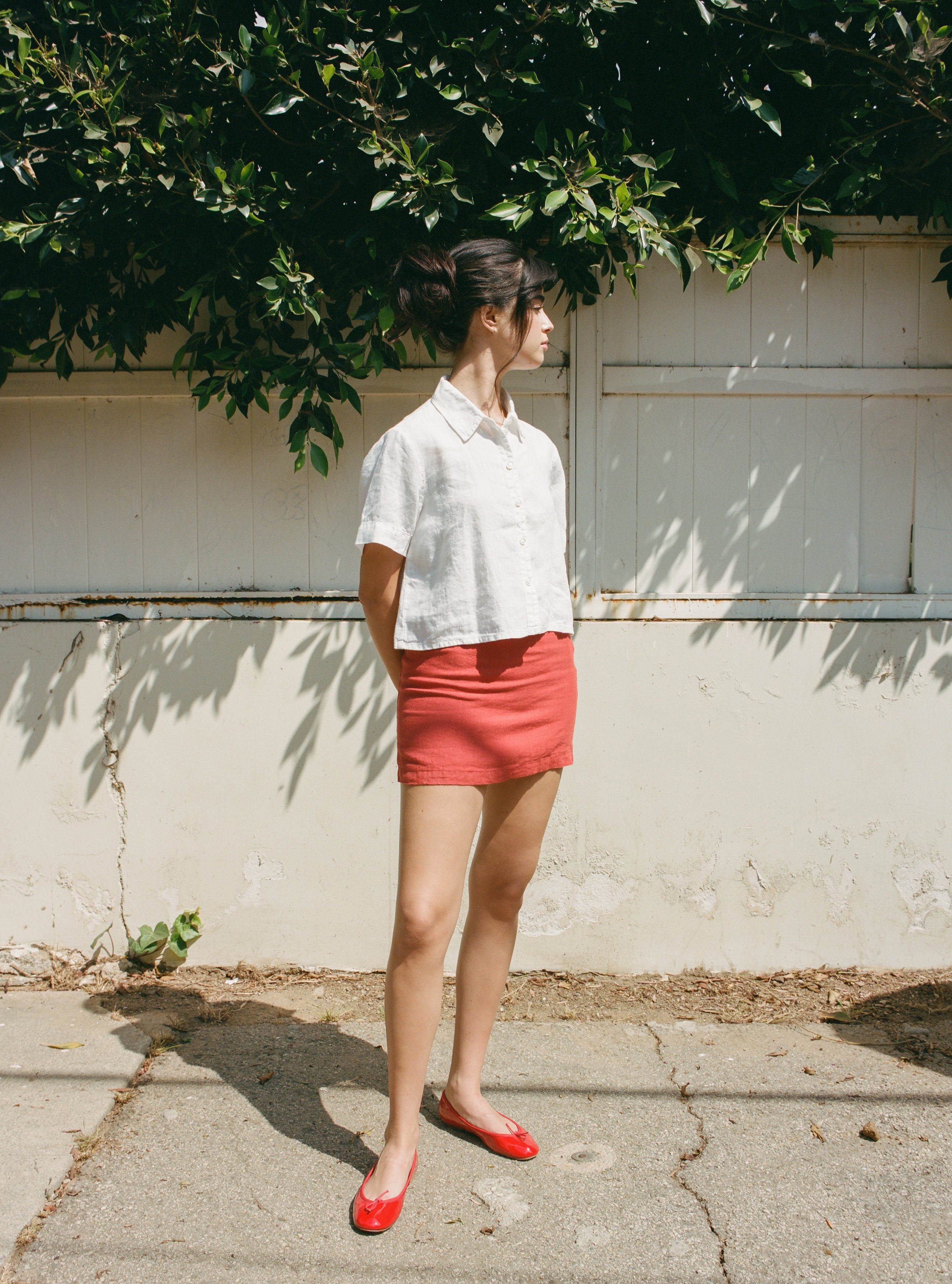 Female model wearing a 100% linen mini skirt with an A-line silhouette in red paired with a white button up short sleeve linen shirt and red flats - VIR VITA
#color_apple red