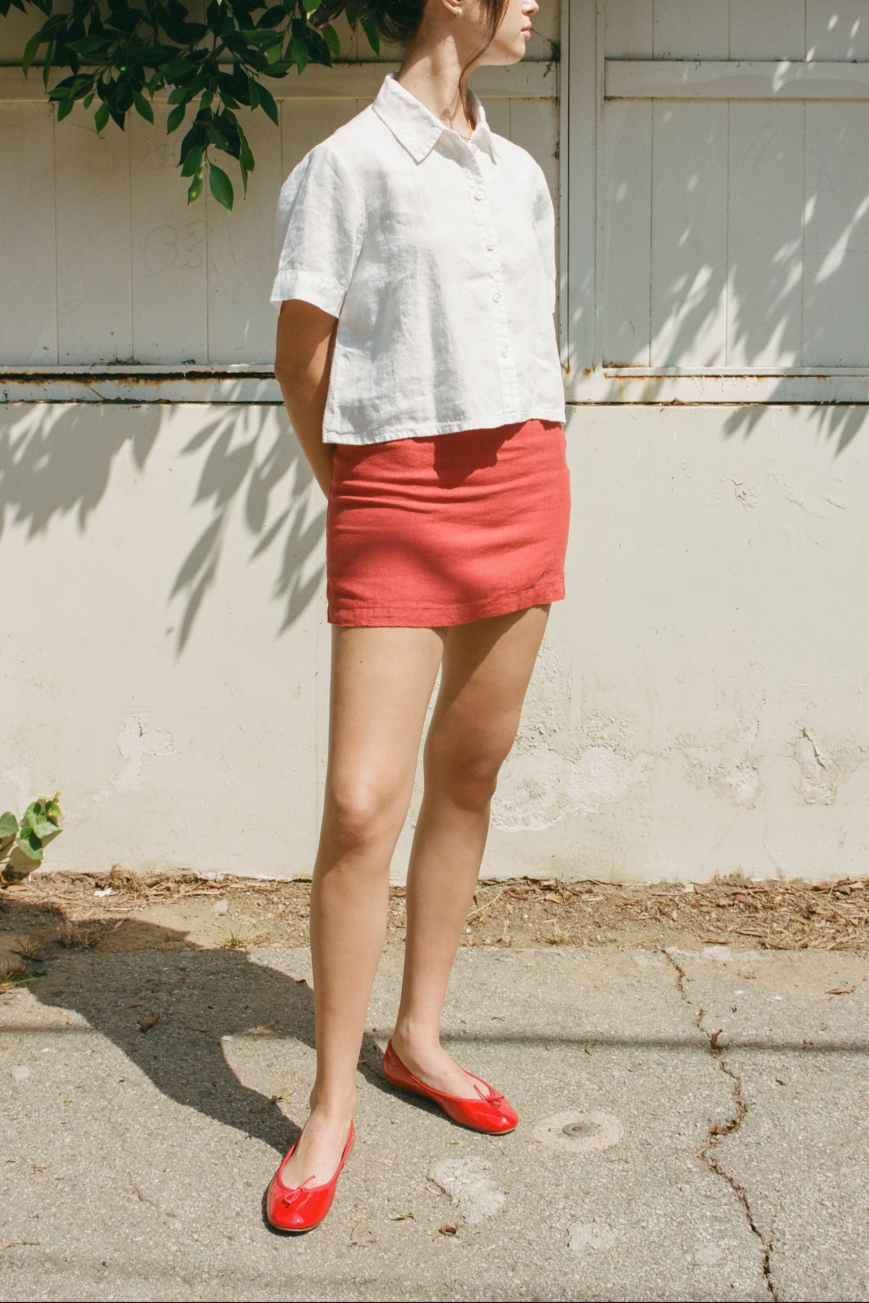 Female model wearing a 100% linen mini skirt with an A-line silhouette in red paired with a white button up short sleeve linen shirt and red flats - VIR VITA
#color_apple red