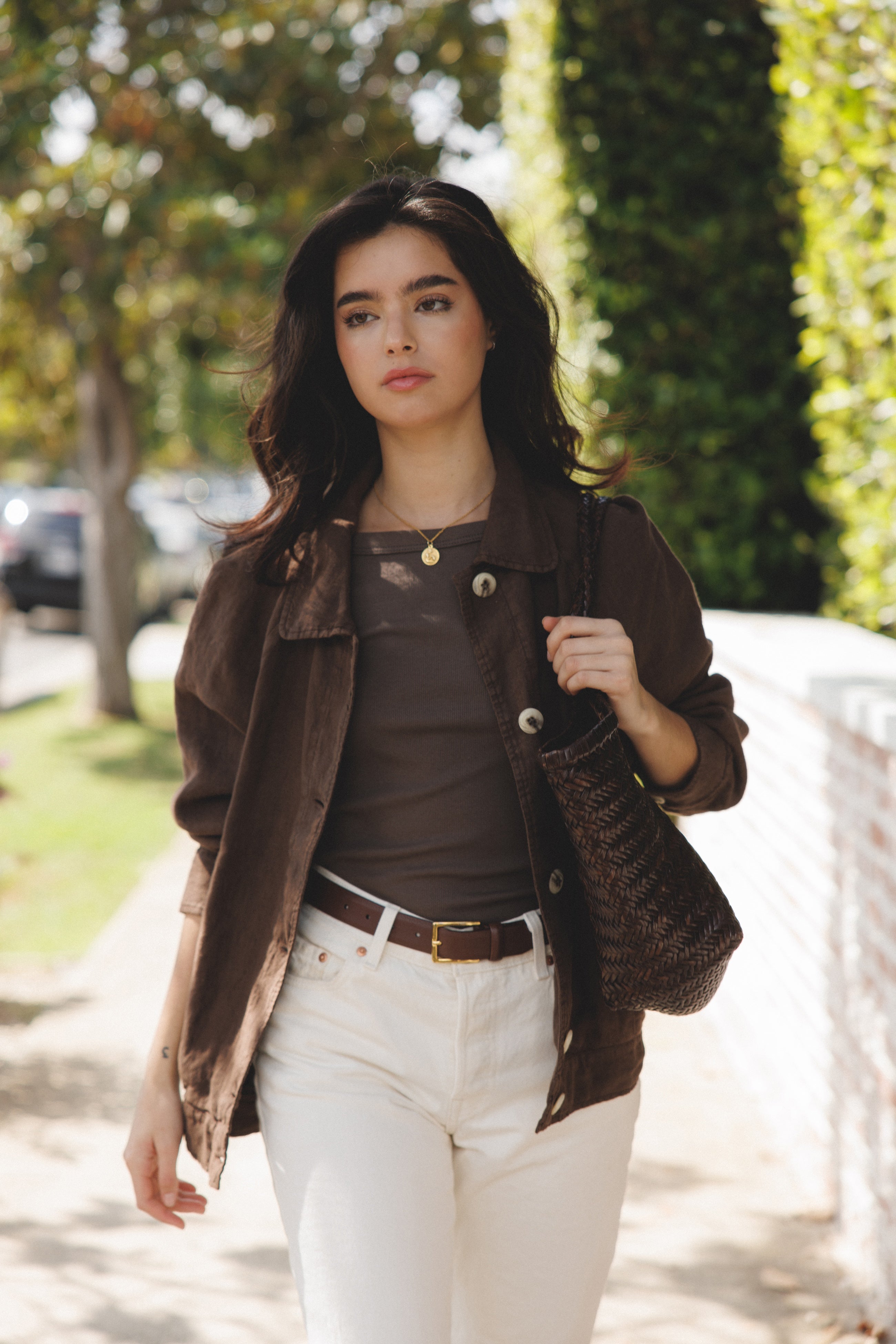 Female model wearing sustainable brown 100% linen barn jacket with brown tank top, white jeans and brown leather belt holding a brown woven bag- VIR VITA
#color_chestnut