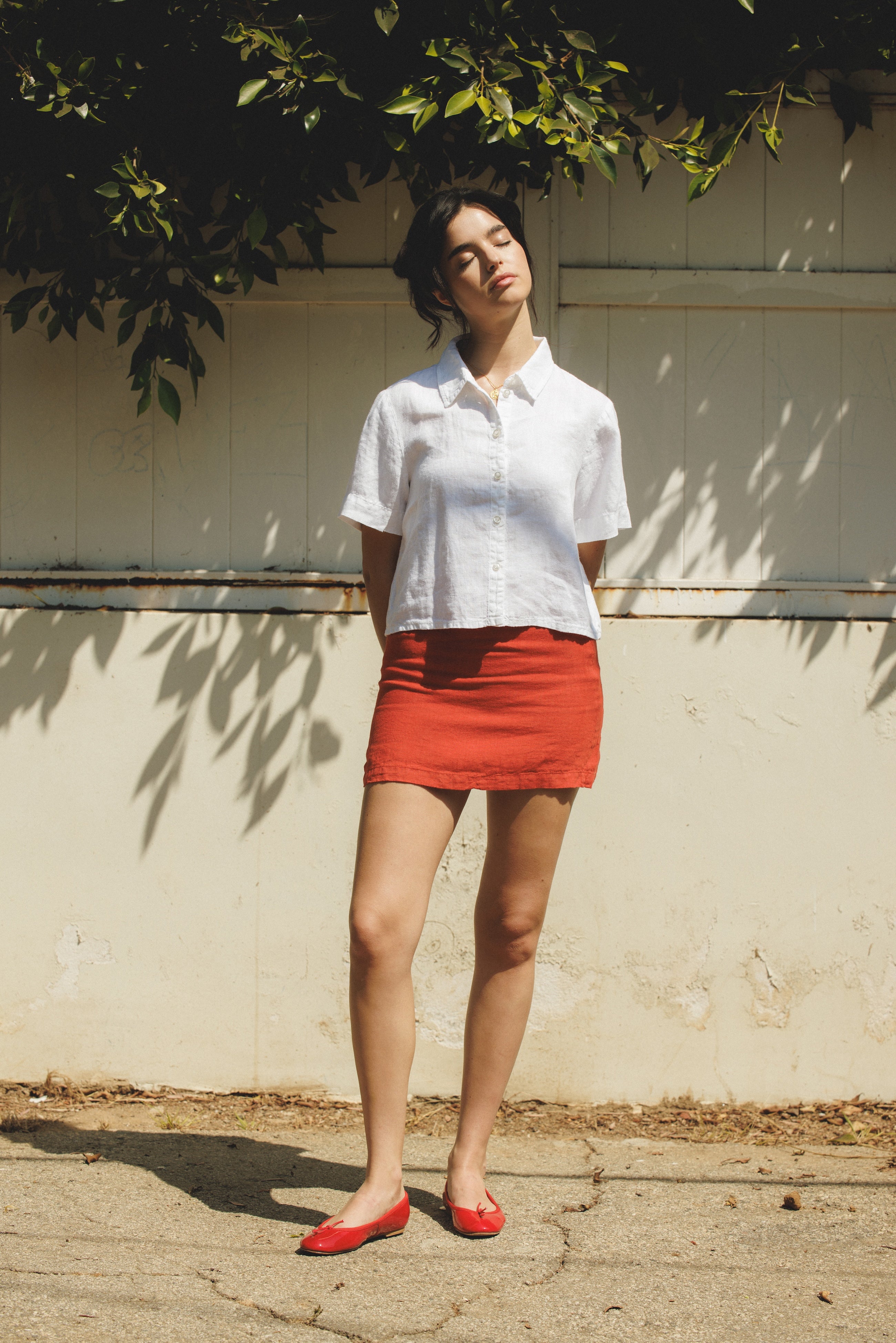 Female model wearing a 100% linen mini skirt with an A-line silhouette in red paired with a white button up short sleeve linen shirt and red flats- VIR VITA
#color_apple red