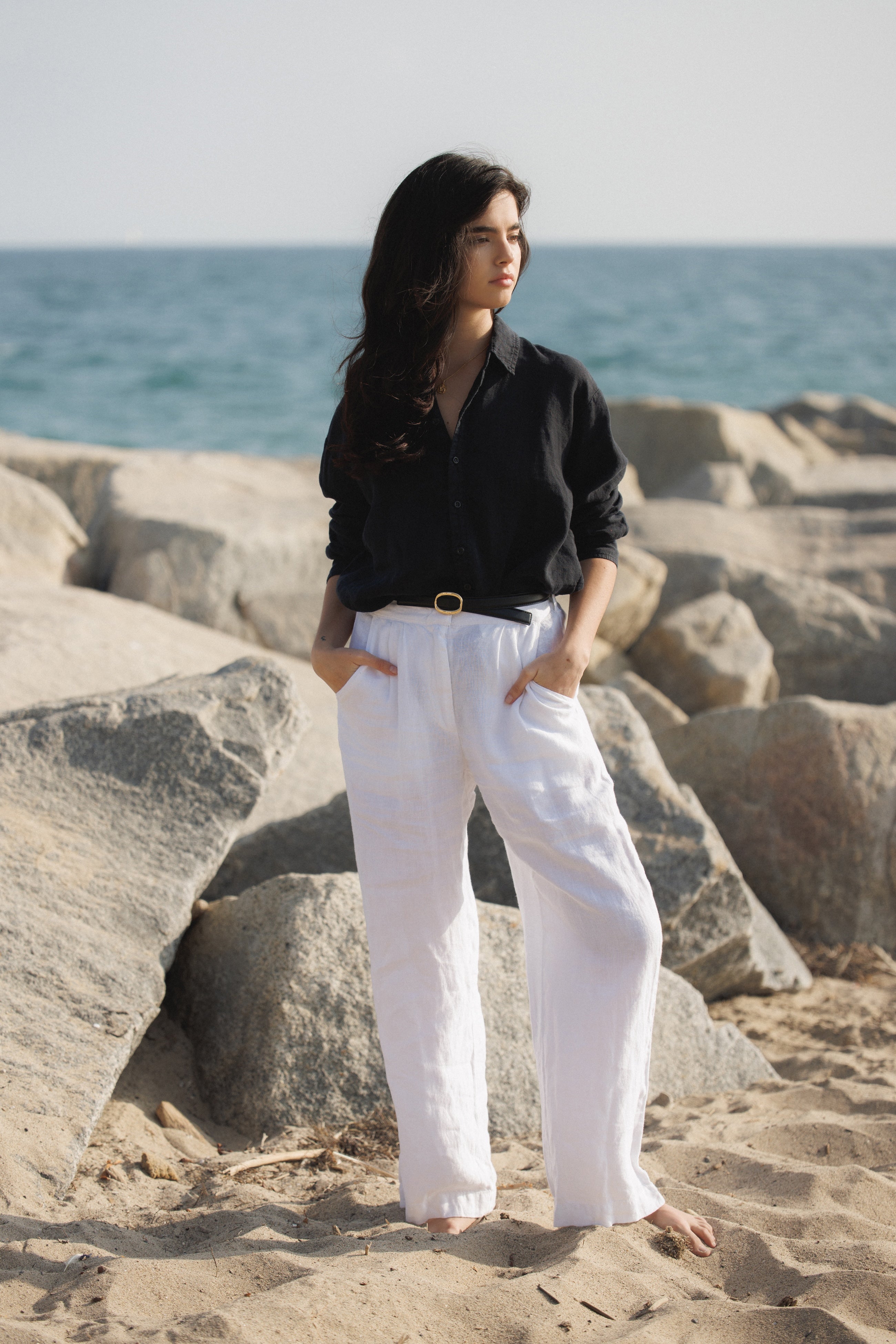Female model wearing sustainable 100% linen oversized button down long sleeve in black paired with 100% linen trousers in white featuring a high waisted double pleat silhouette and a black belt on the beach- VIR VITA
#color_white