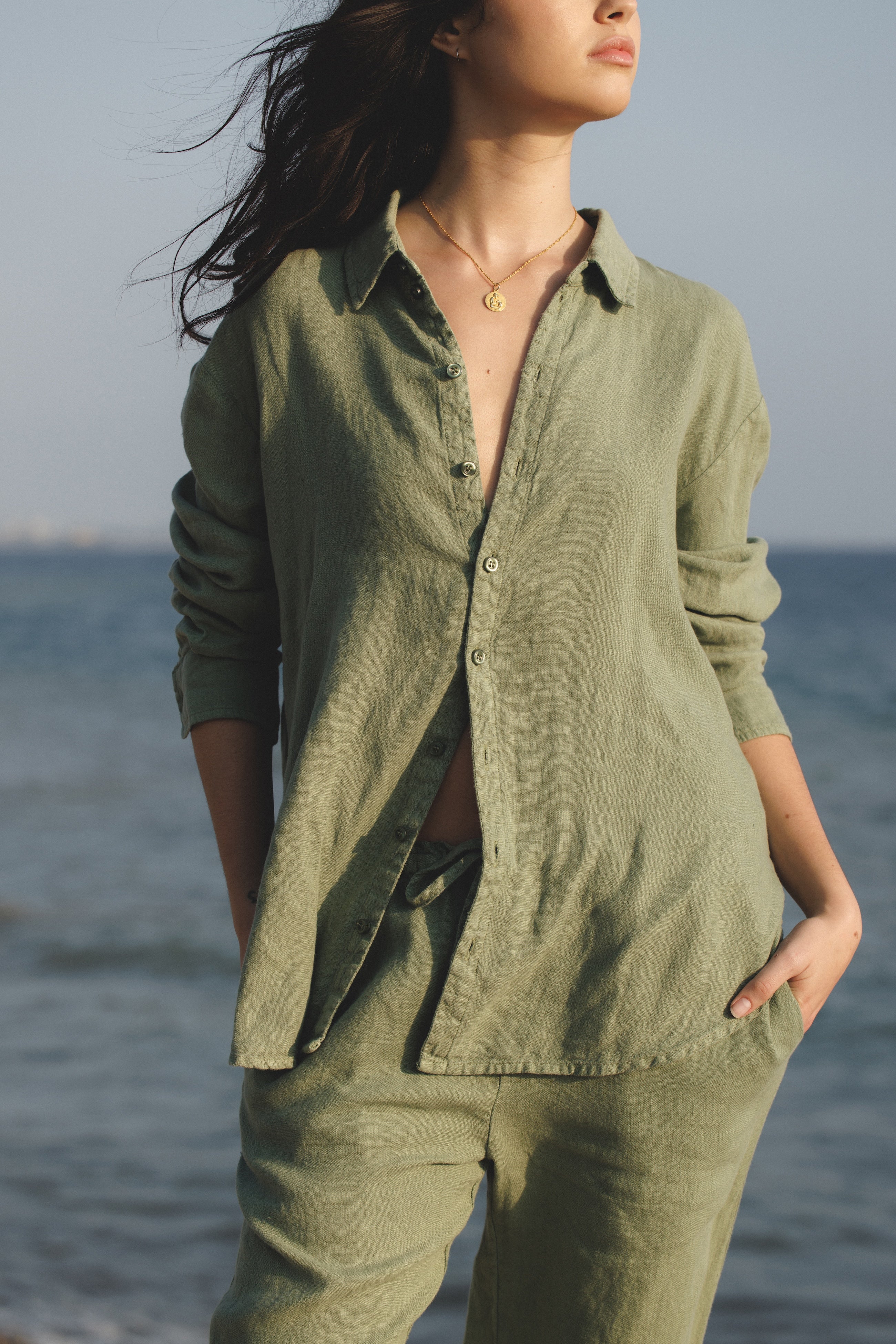 Female model, wearing a sage green colored sustainable oversized 100% linen button down long sleeve with sage green drawstring linen trousers at the beach- VIR VITA
#color_sage