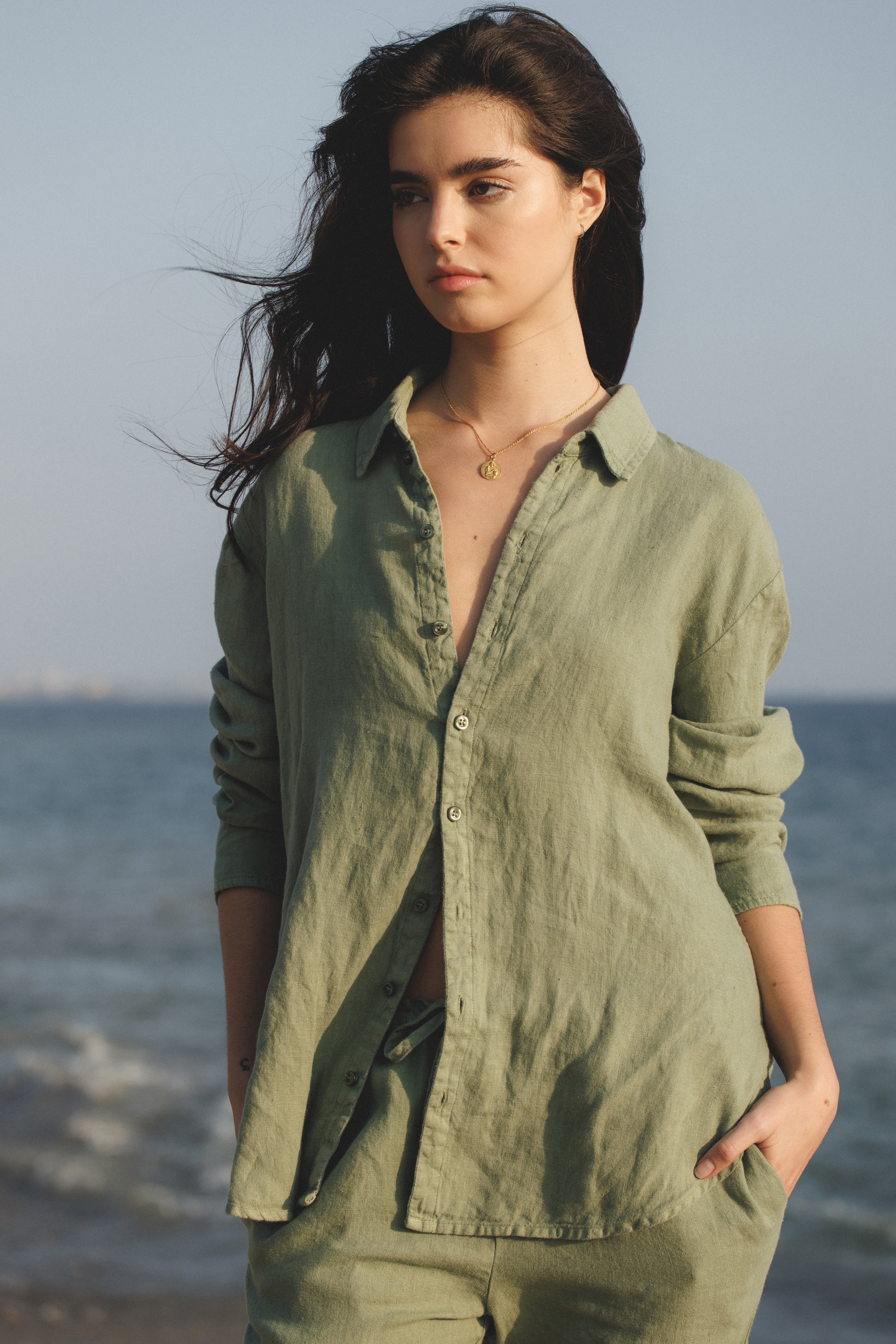 Female model, wearing a sage green colored sustainable oversized 100% linen button down long sleeve with sage green drawstring linen trousers at the beach- VIR VITA
#color_sage