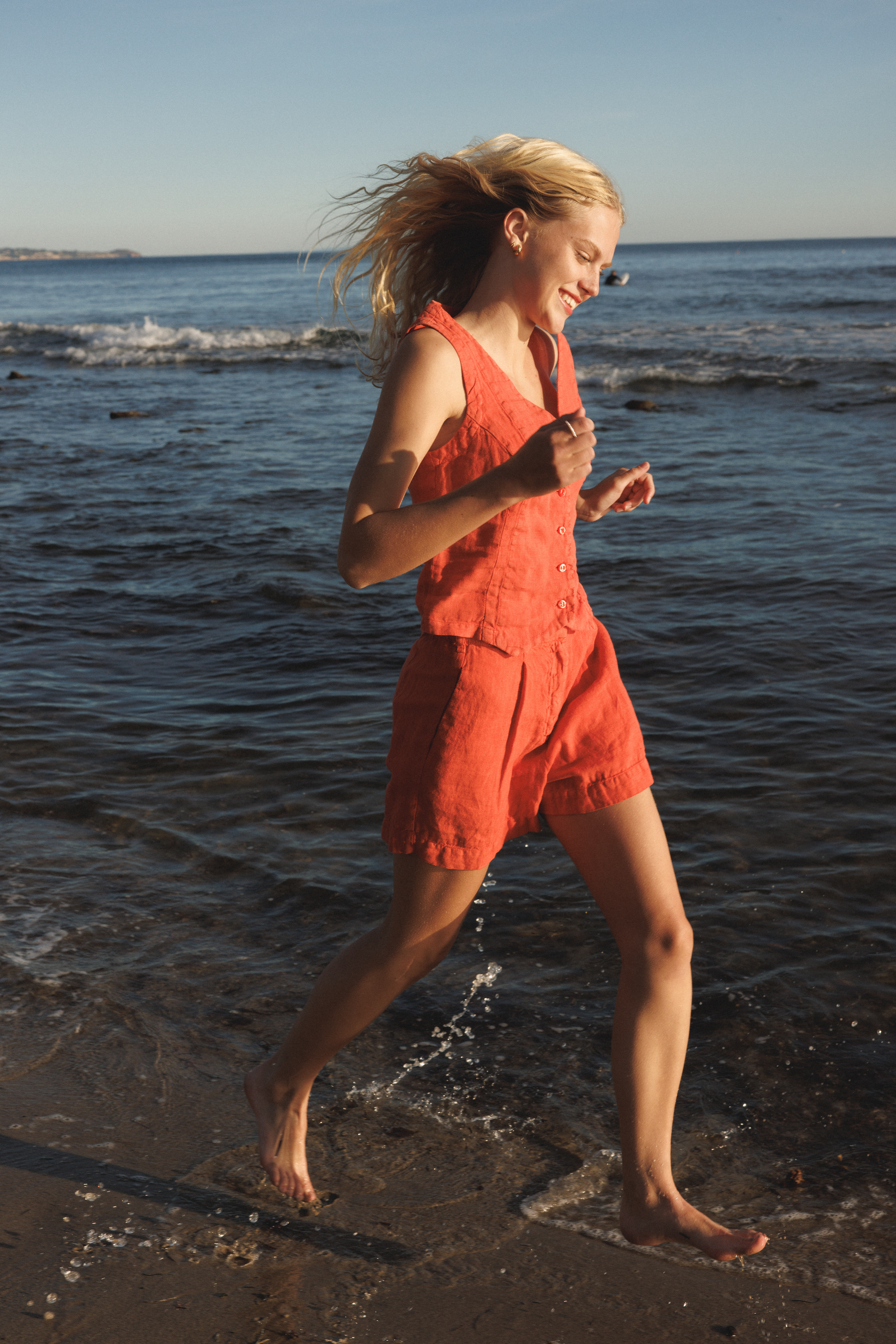 Female model wearing a 100% linen relaxed short featuring double pleats in orange paired with a matching 100% linen vest in orange on the beach - VIR VITA
#color_cayenne