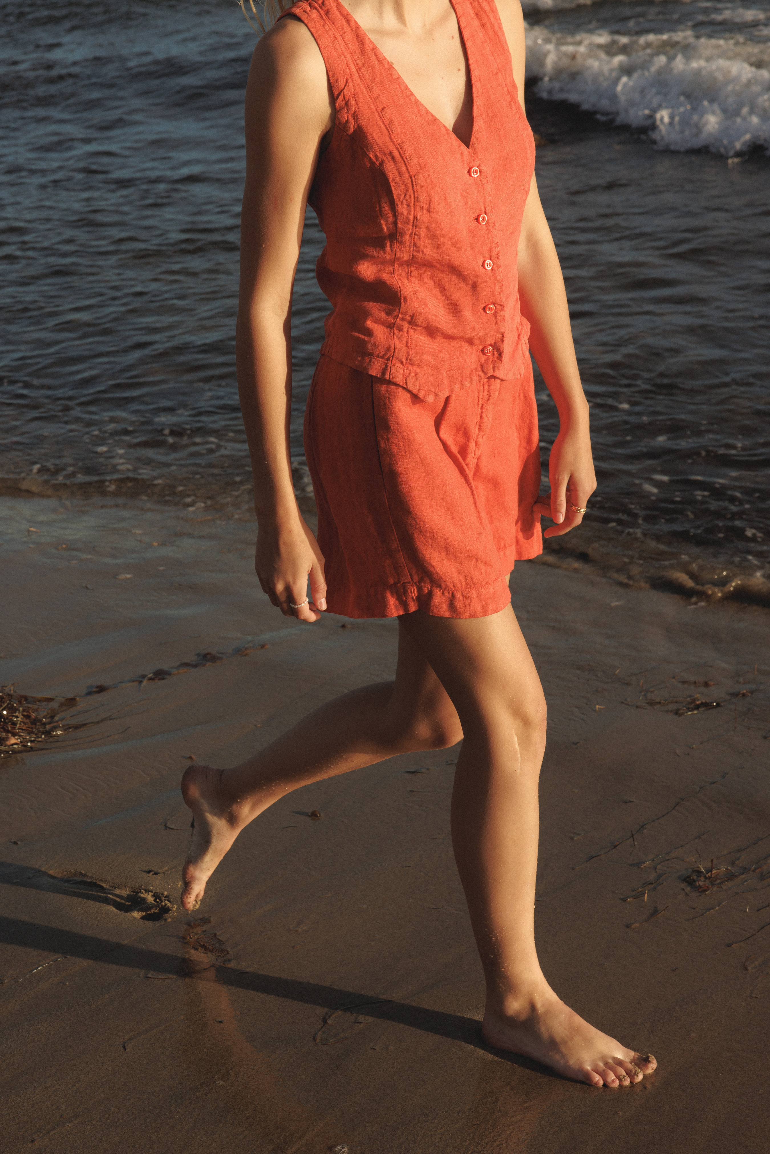 Female model wearing a 100% linen relaxed short featuring double pleats in orange paired with a matching 100% linen vest in orange on the beach - VIR VITA
#color_cayenne