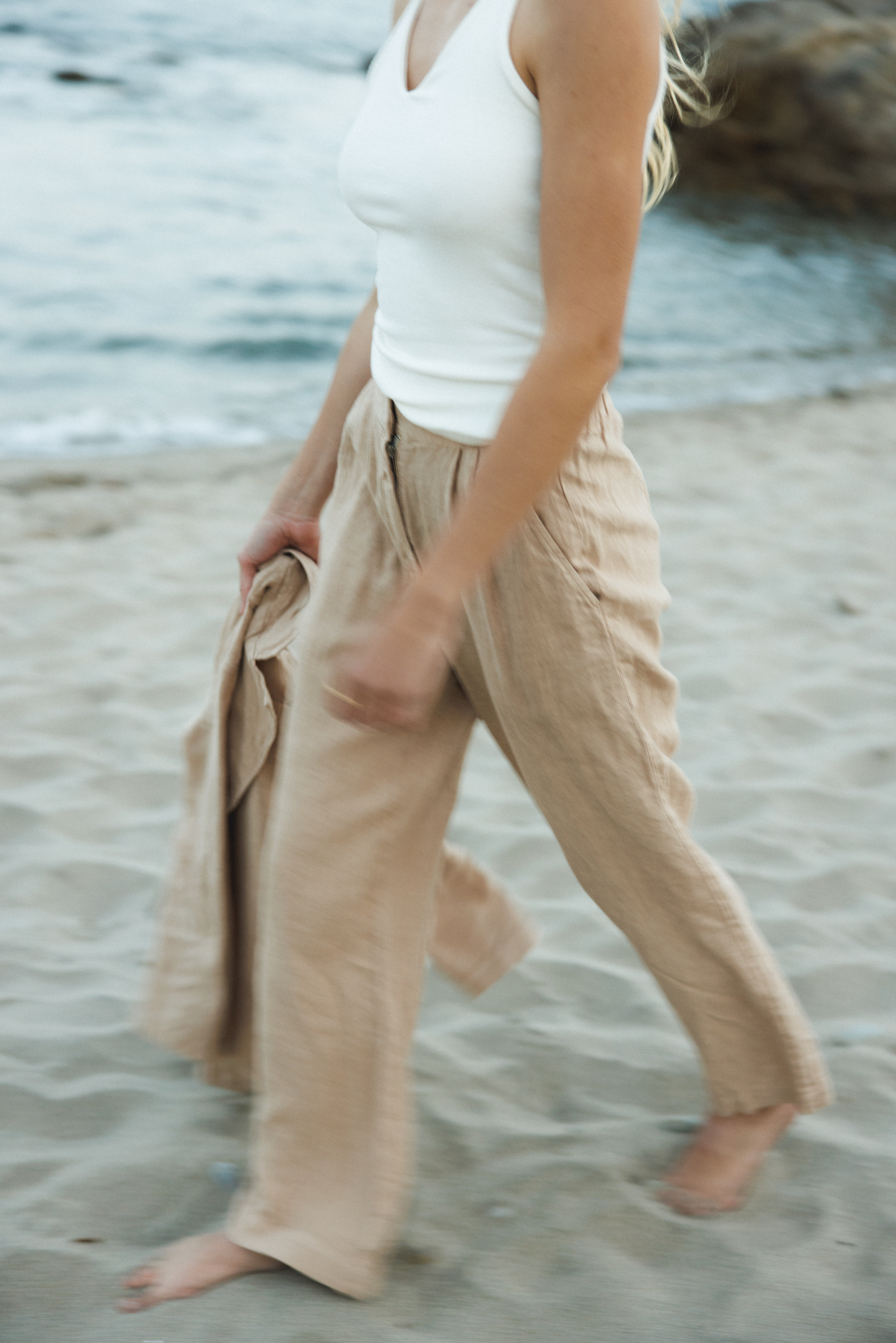 Female model wearing a 100% linen trouser featuring a high waisted double pleat silhouette in beige paired with a 100% linen blazer in beige and a white tank top on the beach- VIR VITA
#color_beige
