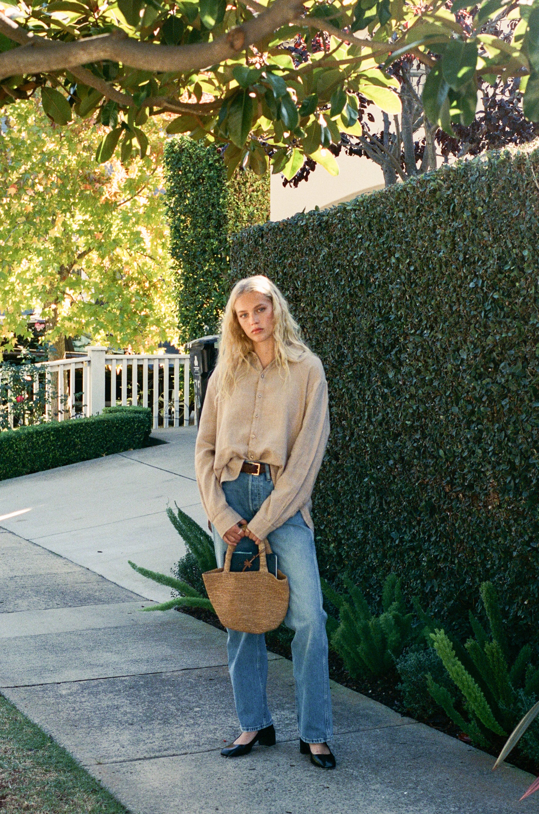 Female model, walking on the sidewalk, wearing a toffee beige sustainable oversized 100% linen button down long sleeve paired with blue jeans, a brown belt, light brown woven bag, and black heels - VIR VITA
#color_toffee