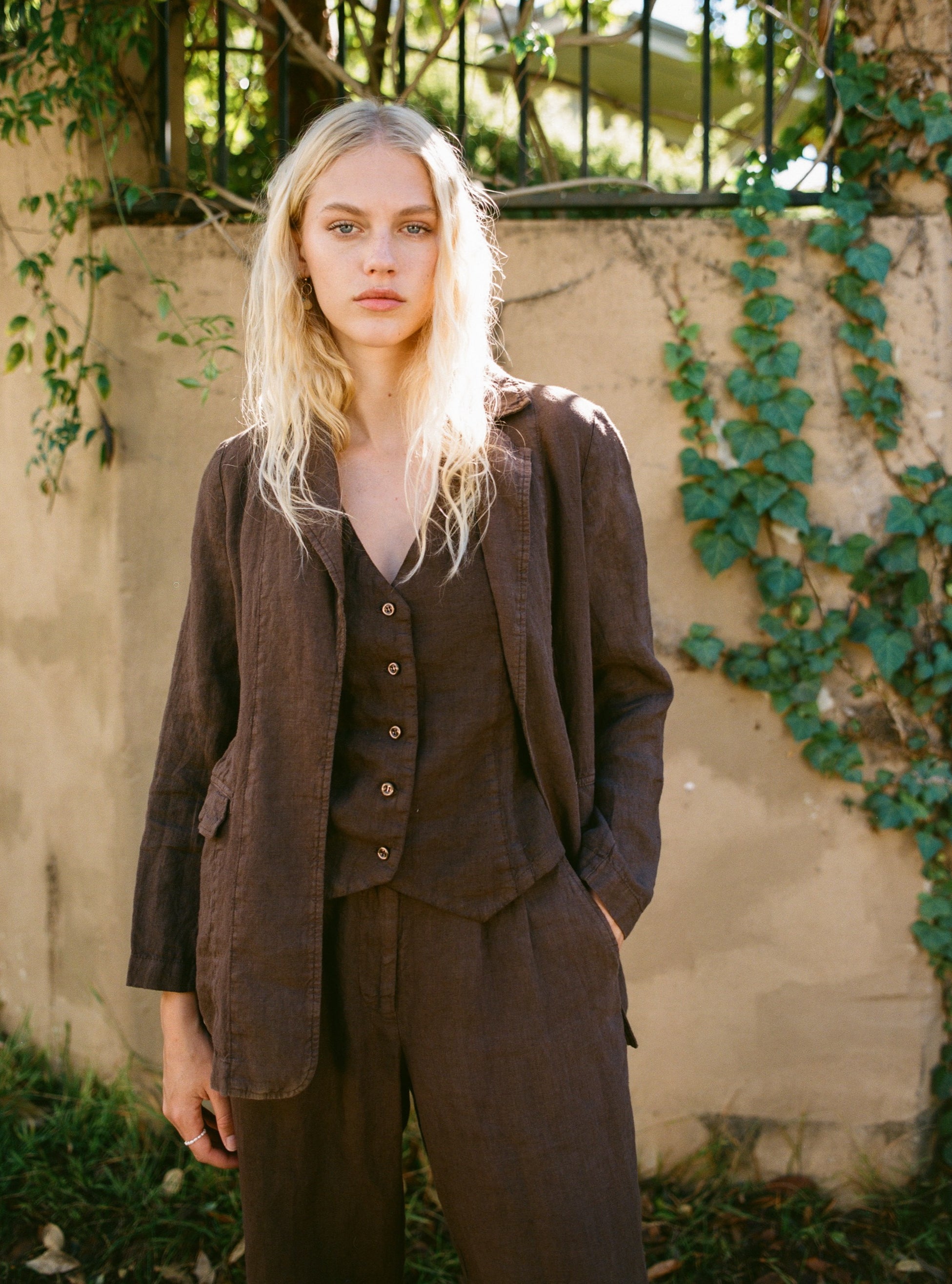 Female model wearing a 100% linen blazer featuring an elegant and classic silhouette in dark brown paired with a matching linen vest in dark brown and linen trousers in dark brown -VIR VITA
#color_chestnut