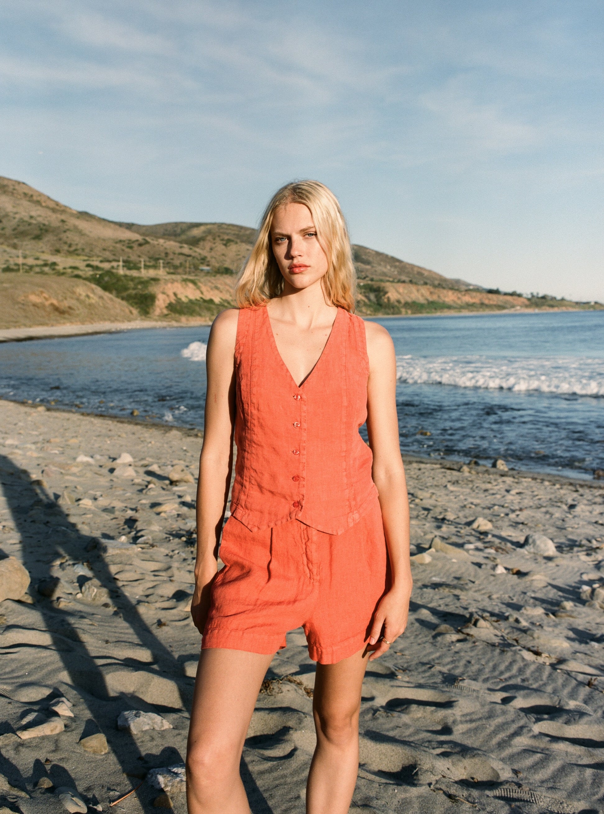 Female model wearing a 100% linen relaxed short featuring double pleats in orange paired with a matching 100% linen vest in orange on the beach - VIR VITA
#color_cayenne