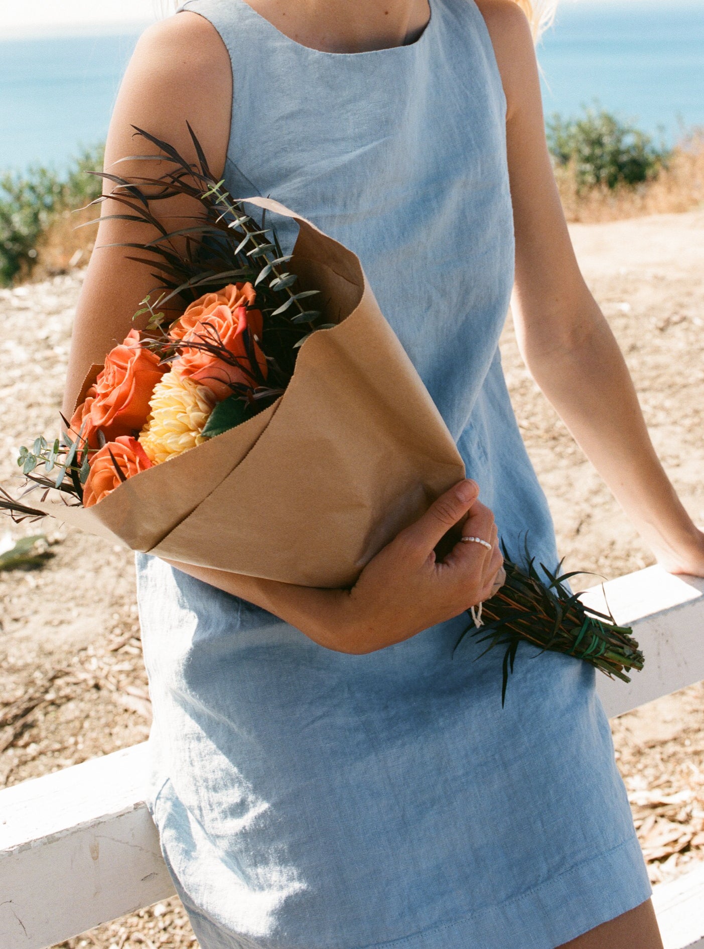 Female model wearing sustainable light blue 100% linen dress with a simple and comfortable silhouette holding a bouquet of flowers overlooking the ocean- VIR VITA
#color_baby blue