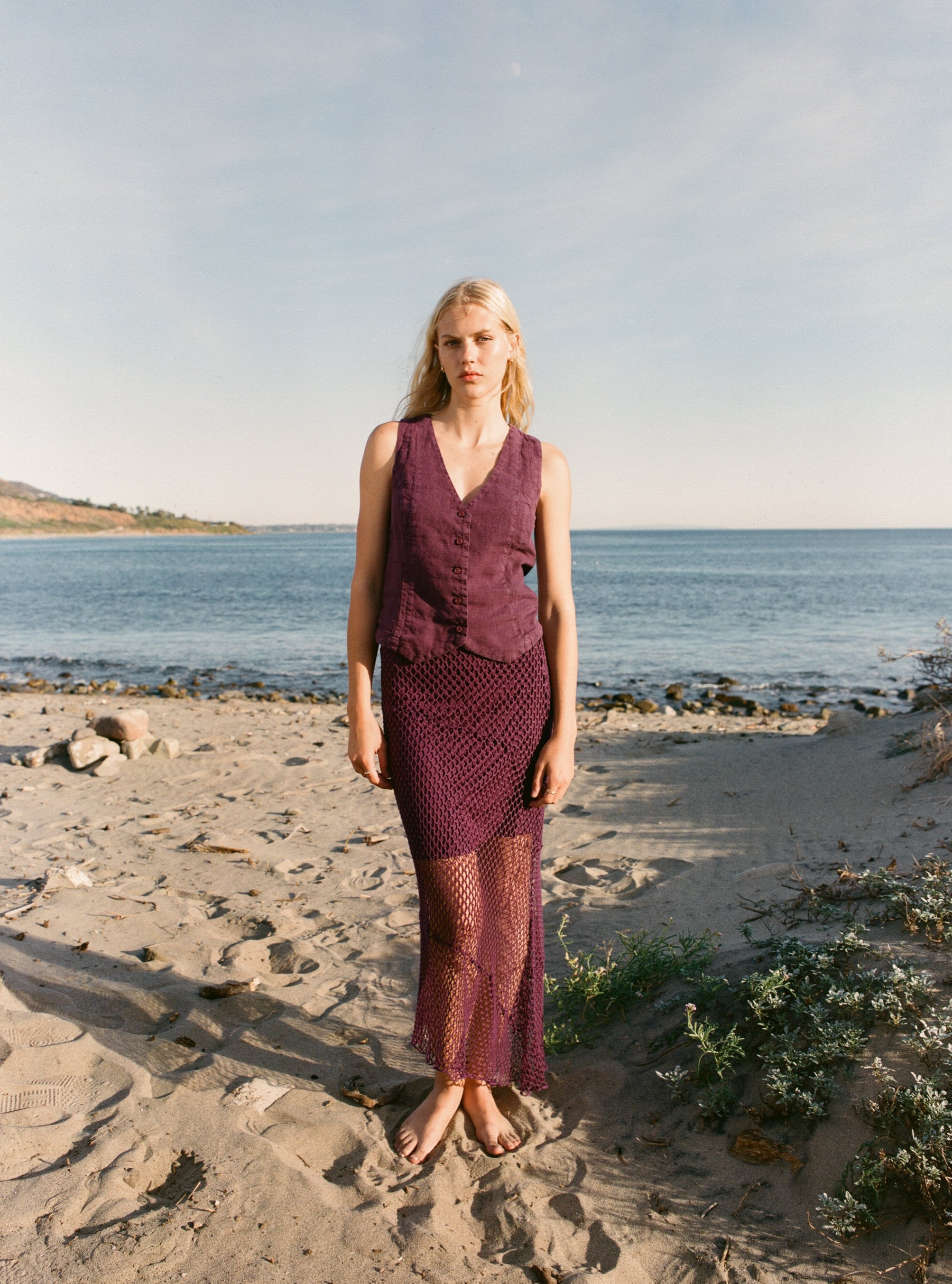 Female model wearing an exquisite crochet skirt featuring an asymmetrical silhouette in plum purple paired with a 100% linen vest in plum purple on the beach - VIR VITA
#color_plum
