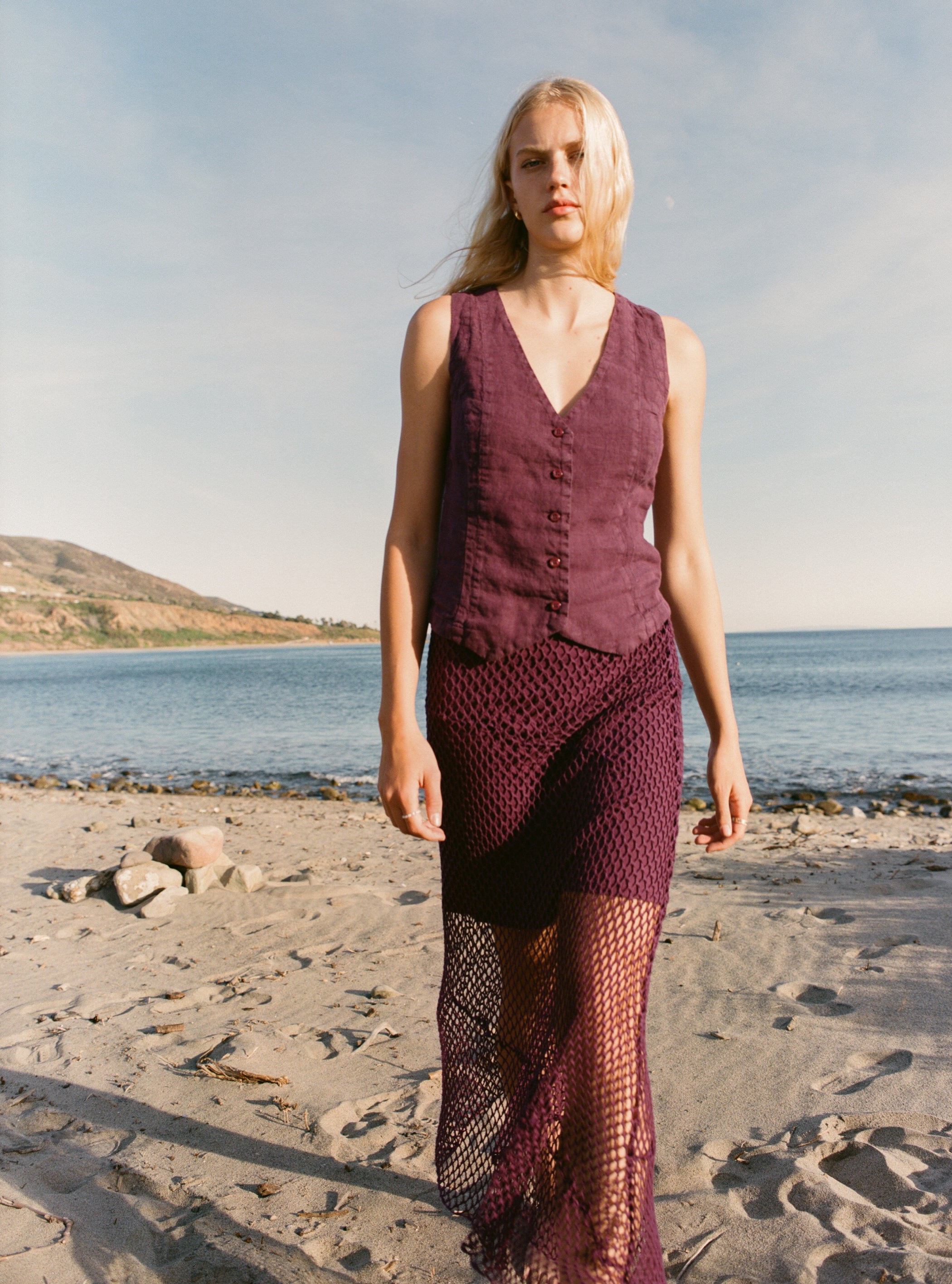 Female model wearing an exquisite crochet skirt featuring an asymmetrical silhouette in plum purple paired with a 100% linen vest in plum purple on the beach - VIR VITA
#color_plum