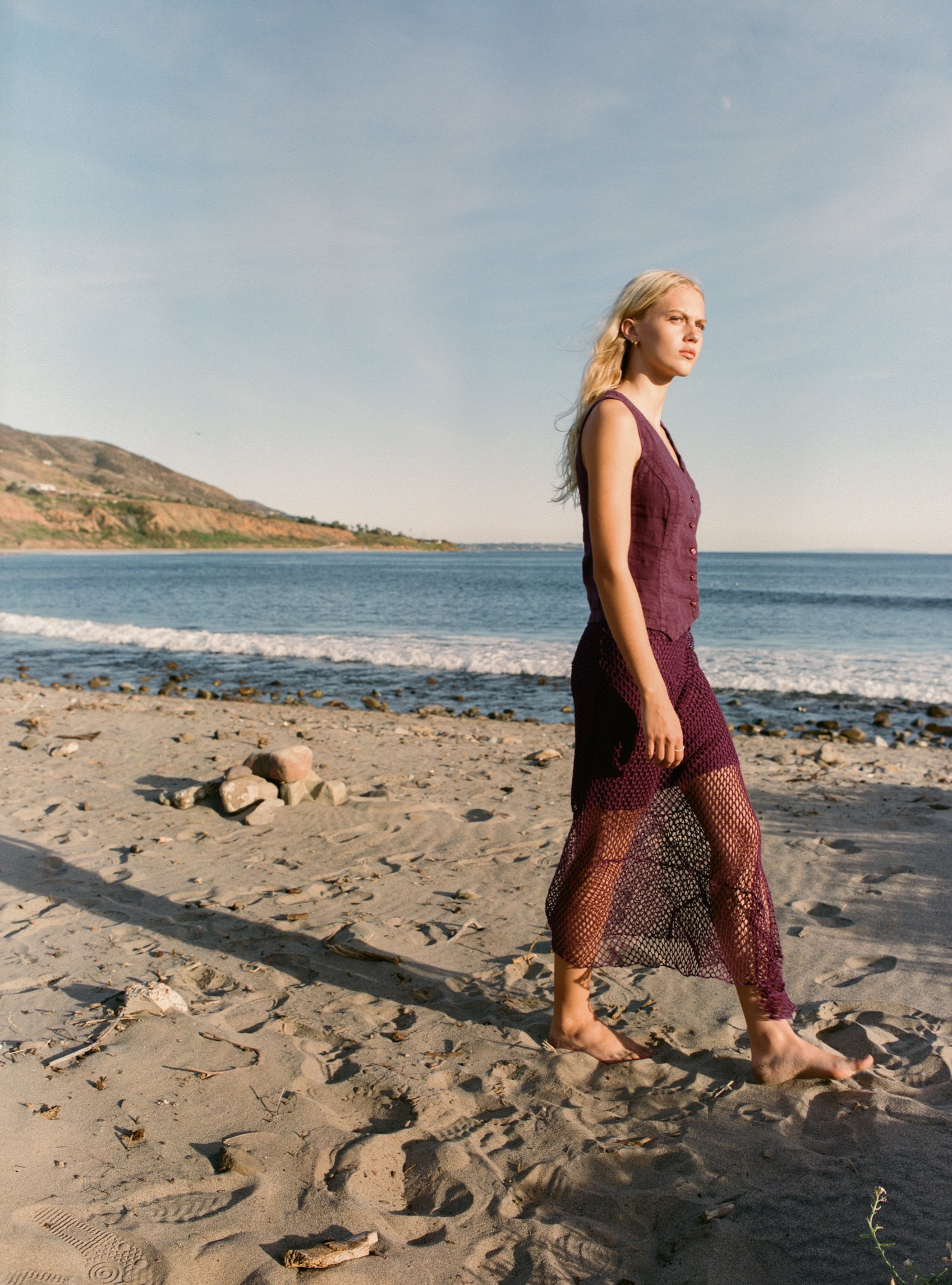 Female model wearing an exquisite crochet skirt featuring an asymmetrical silhouette in plum purple paired with a 100% linen vest in plum purple on the beach - VIR VITA
#color_plum