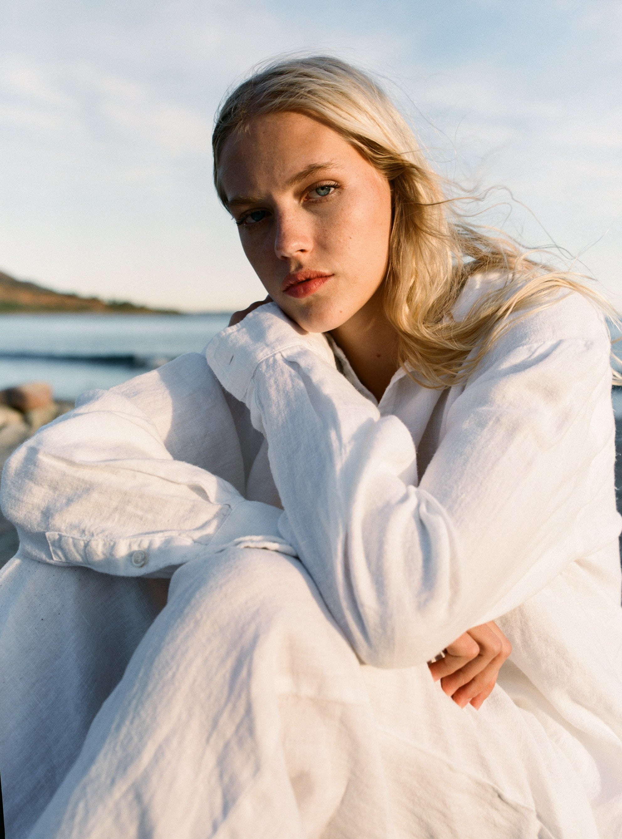 Female model, sitting in front of the ocean, wearing a white sustainable oversized 100% linen button down long sleeve paired with matching white linen pants - VIR VITA
#color_white