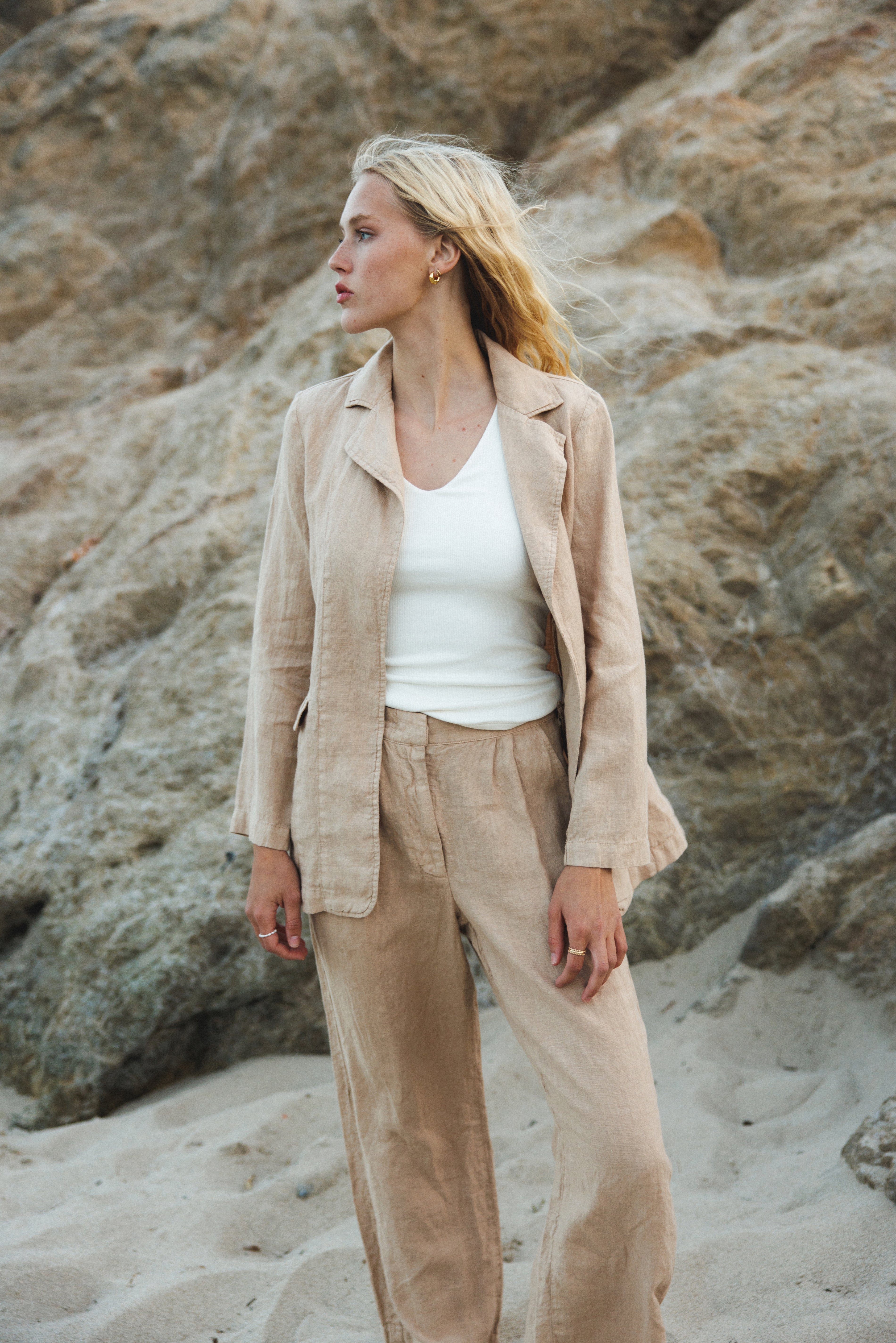 Female model wearing a 100% linen trouser featuring a high waisted double pleat silhouette in beige paired with a 100% linen blazer in beige and a white tank top on the beach - VIR VITA
#color_beige