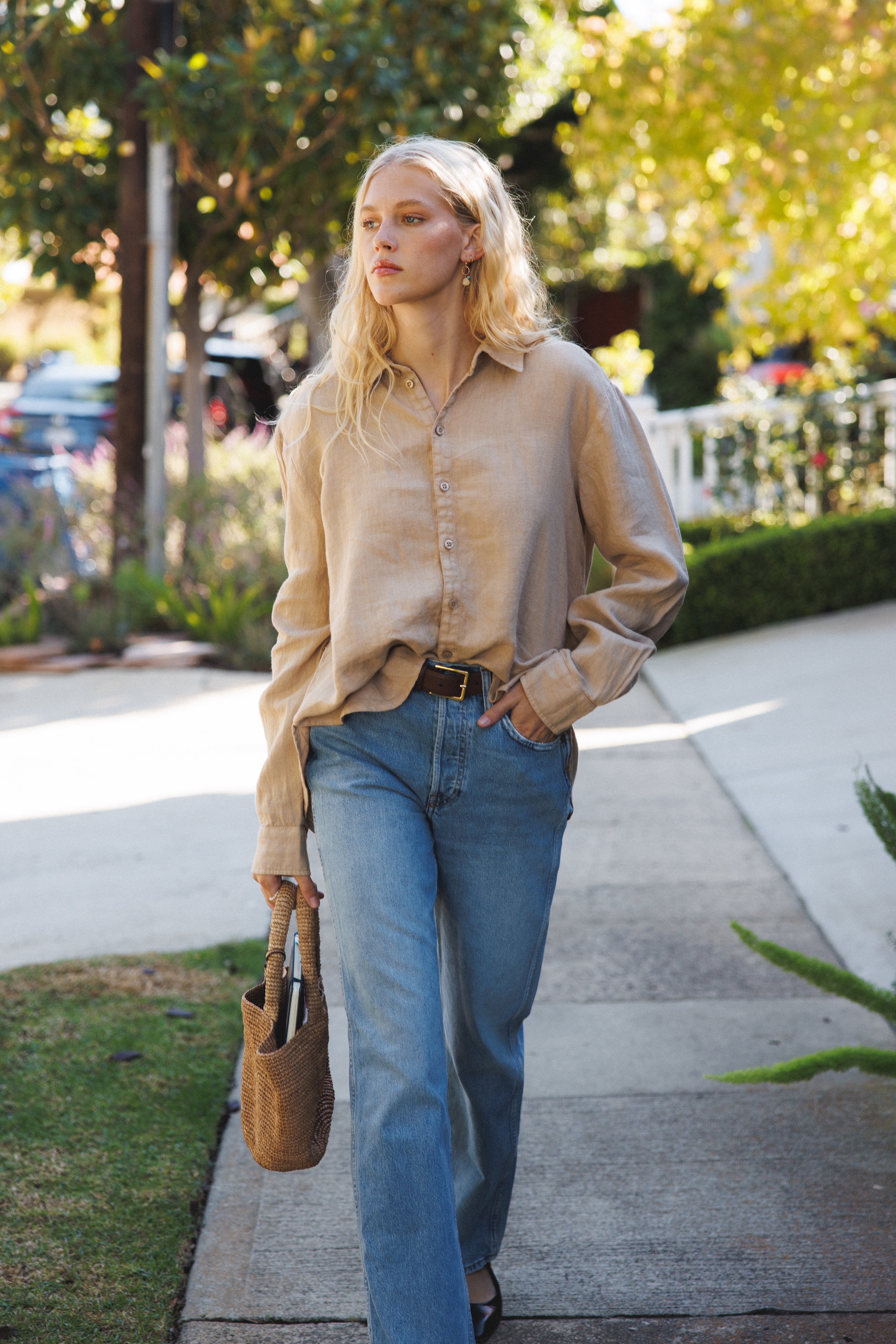 Female model, walking on the street, wearing a toffee beige sustainable oversized 100% linen button down long sleeve paired with blue jeans, a brown belt, and light brown woven bag - VIR VITA
#color_toffee