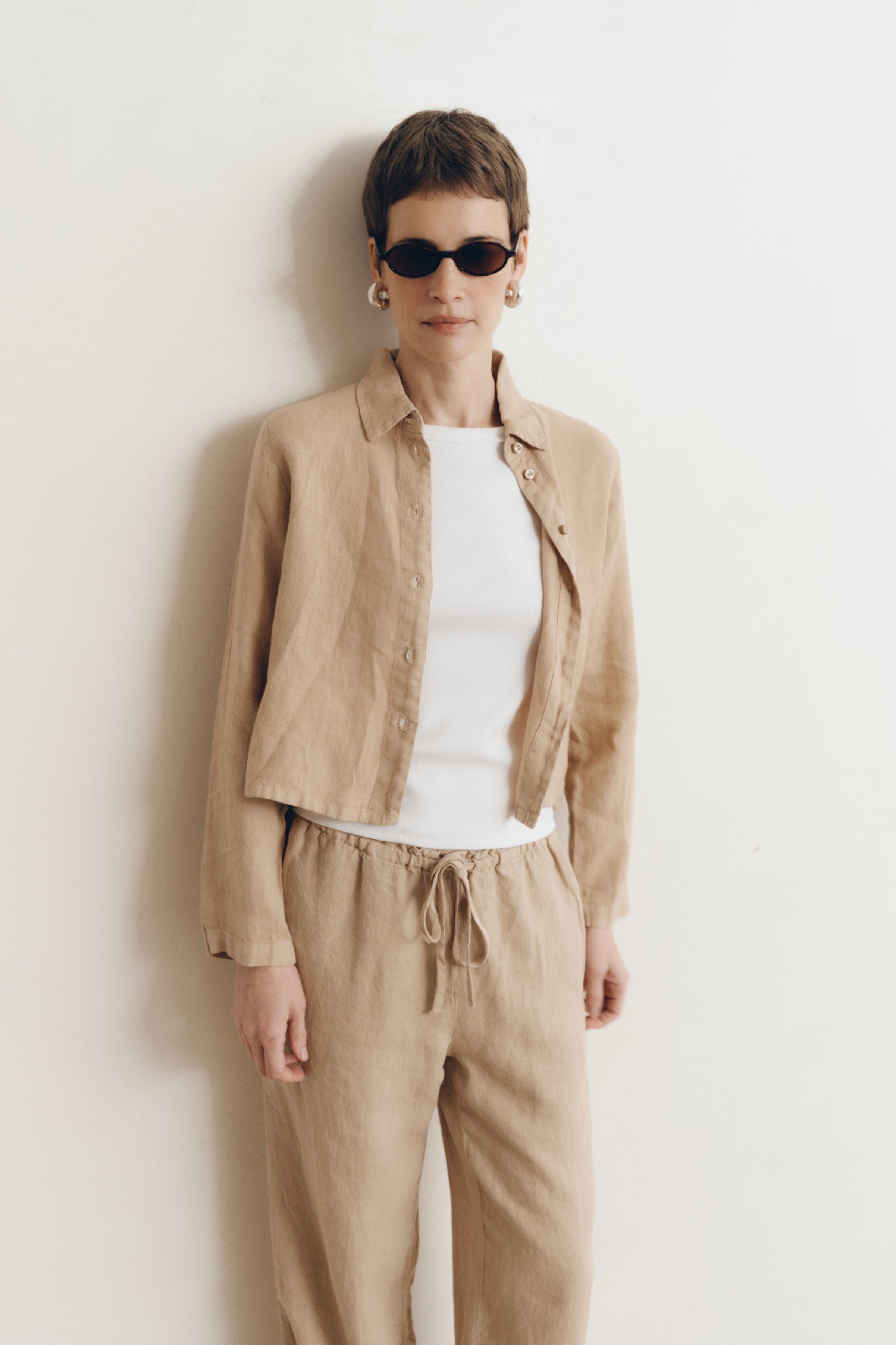 Posed female model wearing a sustainable 100% linen classic button up collared shirt in toffee beige paired with matching drawstring linen pants, a high neck white tank top, and black sunglasses - VIR VITA
#color_toffee