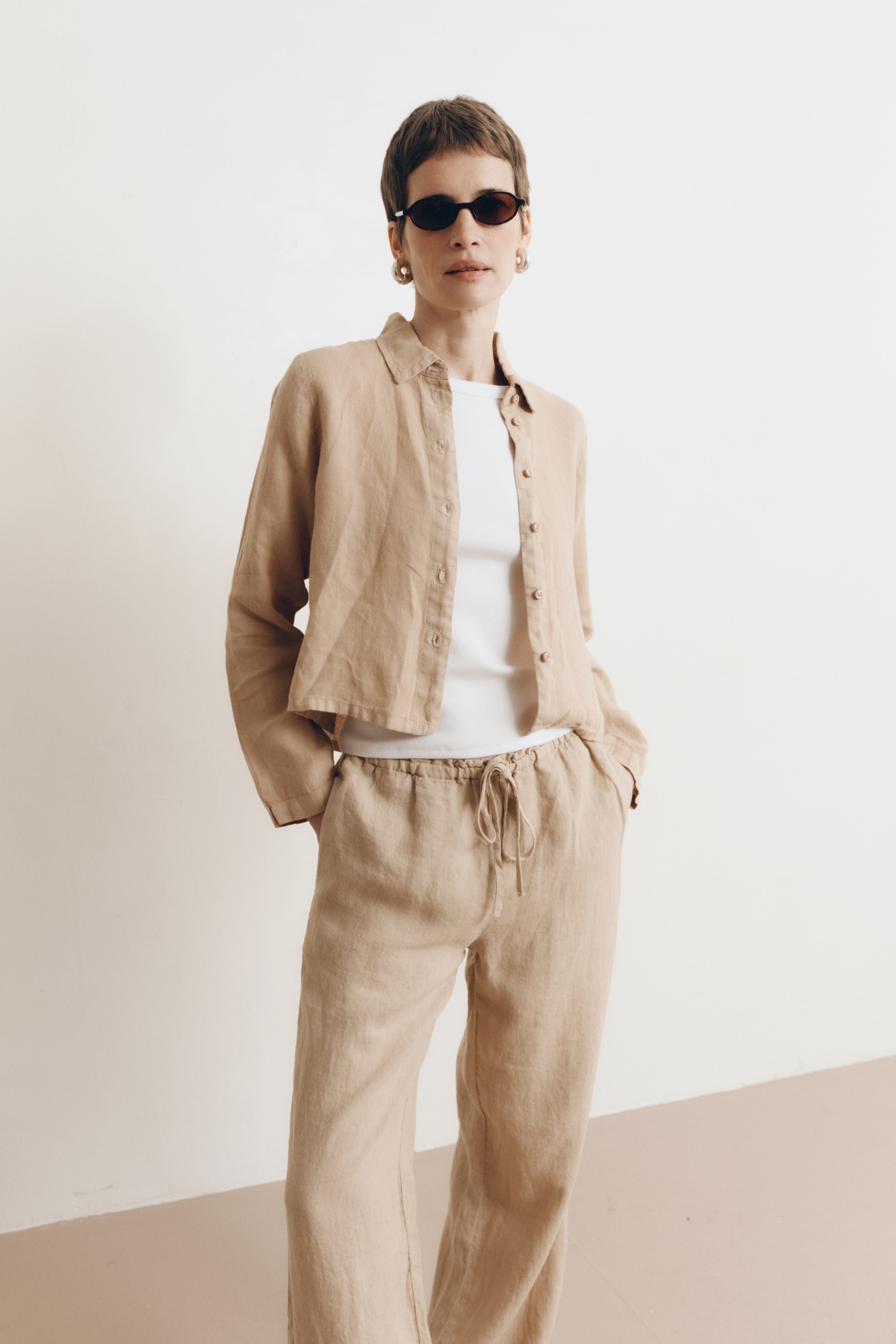 Posed female model wearing a sustainable 100% linen classic button up collared shirt in toffee beige paired with matching drawstring linen pants, a high neck white tank top, and black sunglasses - VIR VITA
#color_toffee