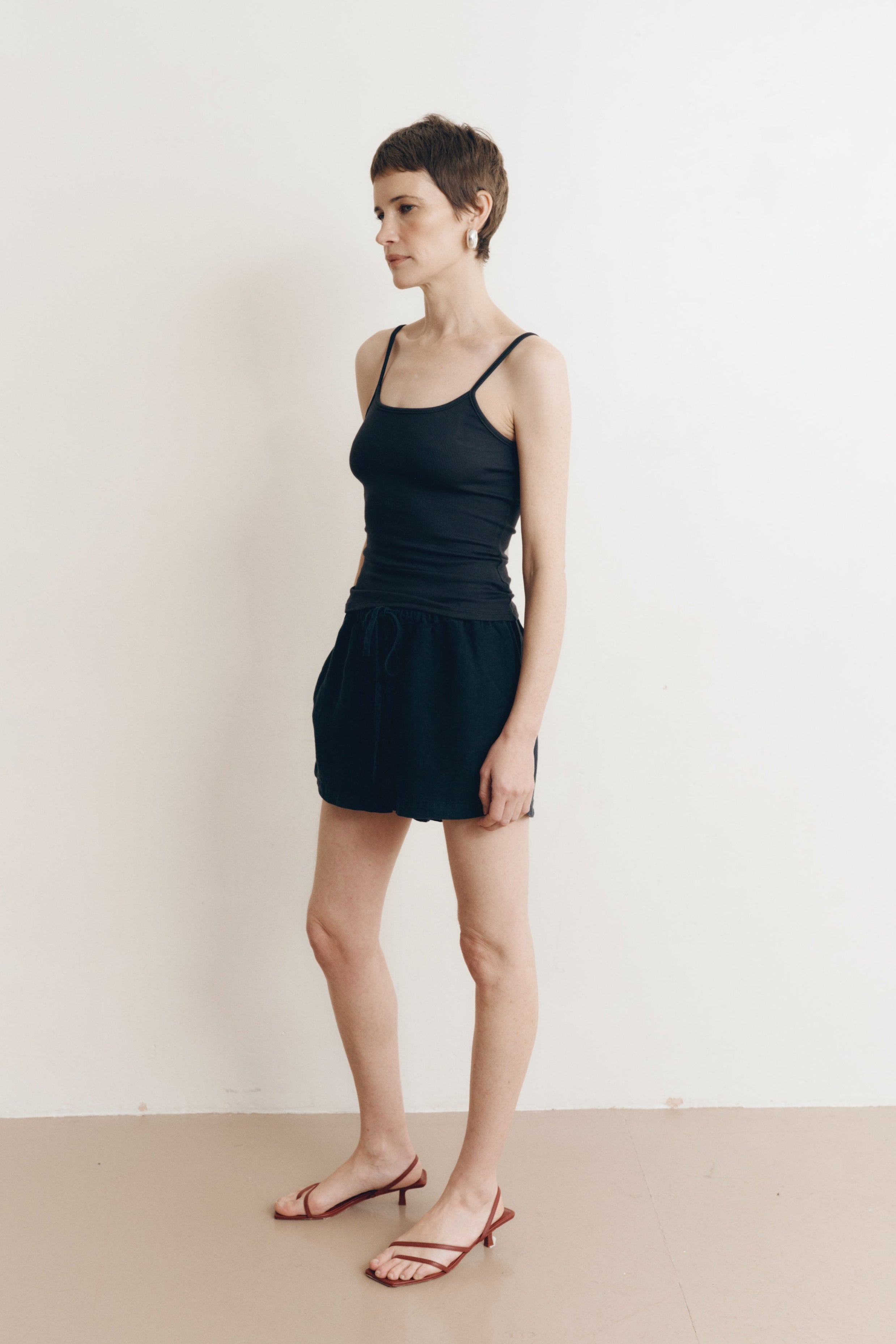 Posed female model wearing sustainable 100% linen drawstring shorts in black paired with a modal cami tank top in black, and brown heels - VIR VITA
#color_black