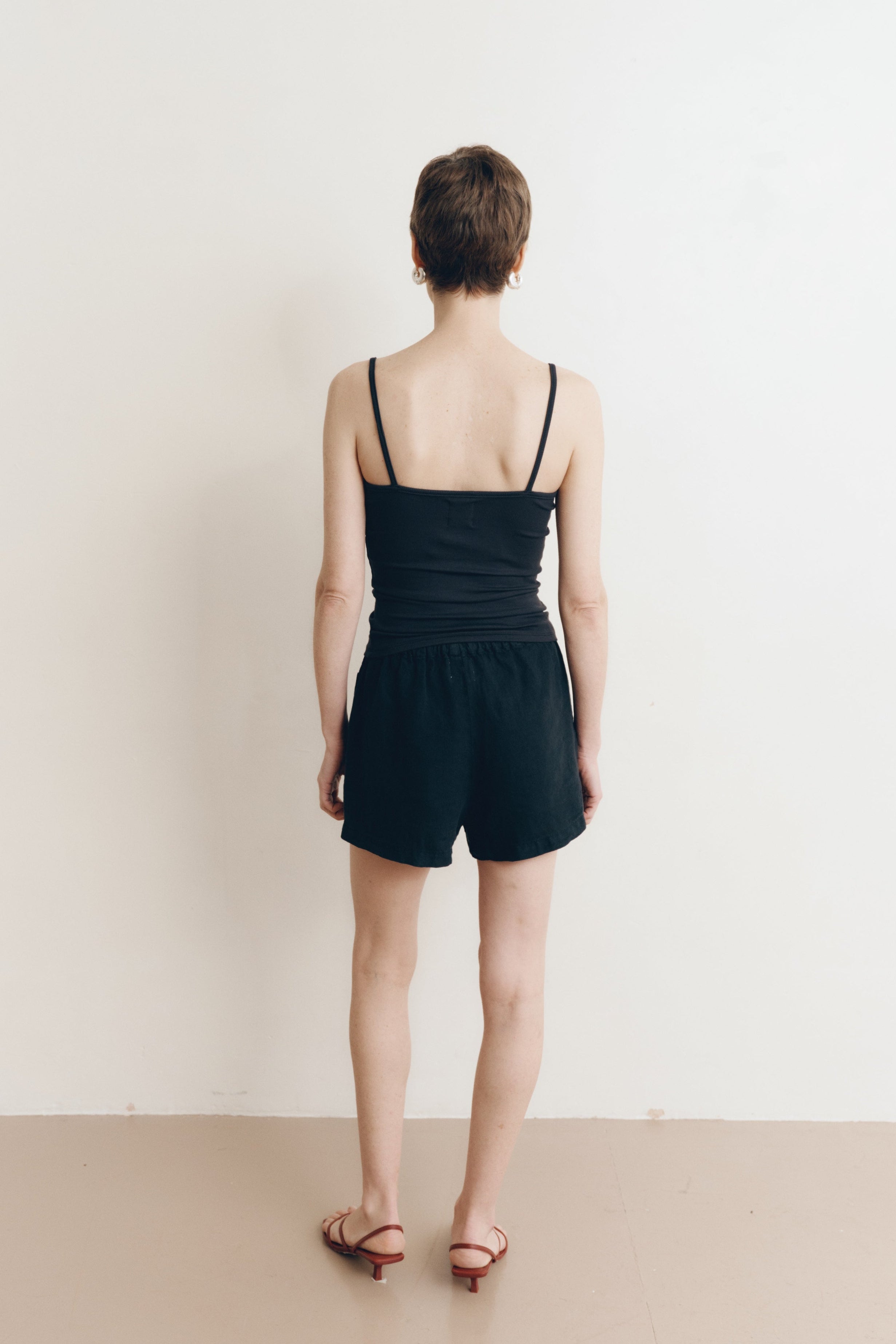 Posed female model wearing sustainable 100% linen drawstring shorts in black paired with a modal cami tank top in black, and brown heels - VIR VITA
#color_black