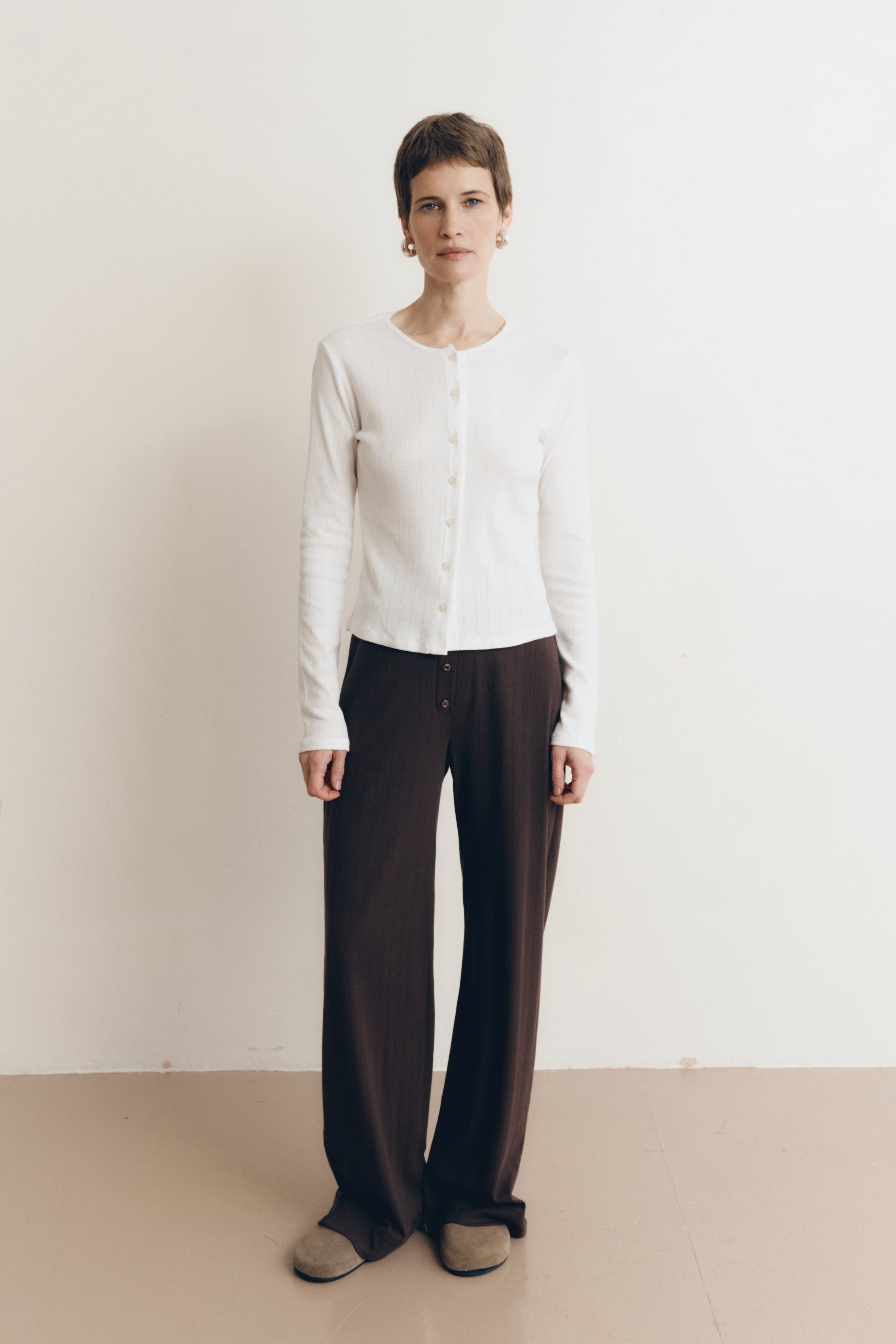 Posed female model wearing a button up sustainable 100% cotton pointelle knit cardigan in white paired with pointelle lounge pants in dark brown and brown suede shoes- VIR VITA
#color_coconut